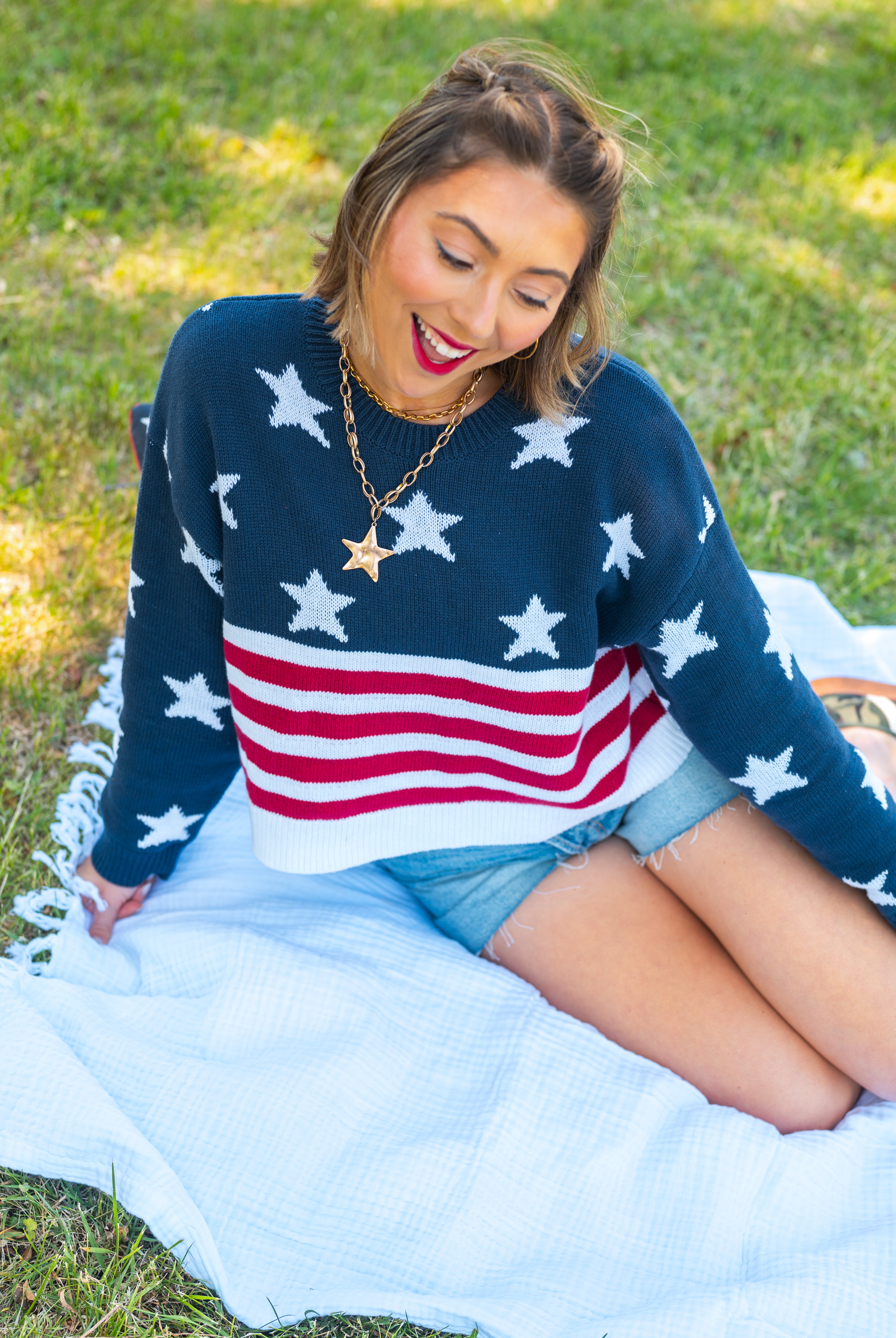 Woman wearing a sweater with an American flag design, sitting on grass, USA, cropped sweater, Americana, red white and blue, chunky sweater, stars and stripes