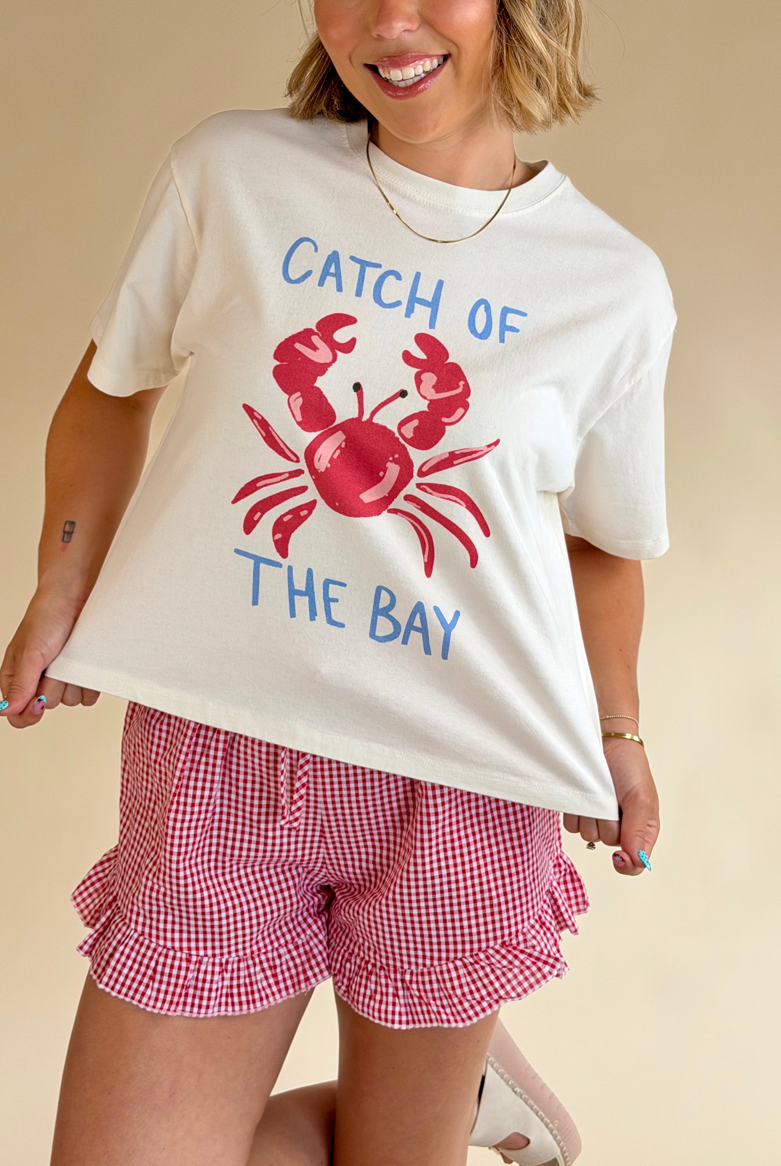 Person wearing a white t-shirt with a red crab graphic and text, paired with red checkered shorts on a beige background, crab graphic tee, cropped fit tee, red white and blue, summer staple, crab graphic
