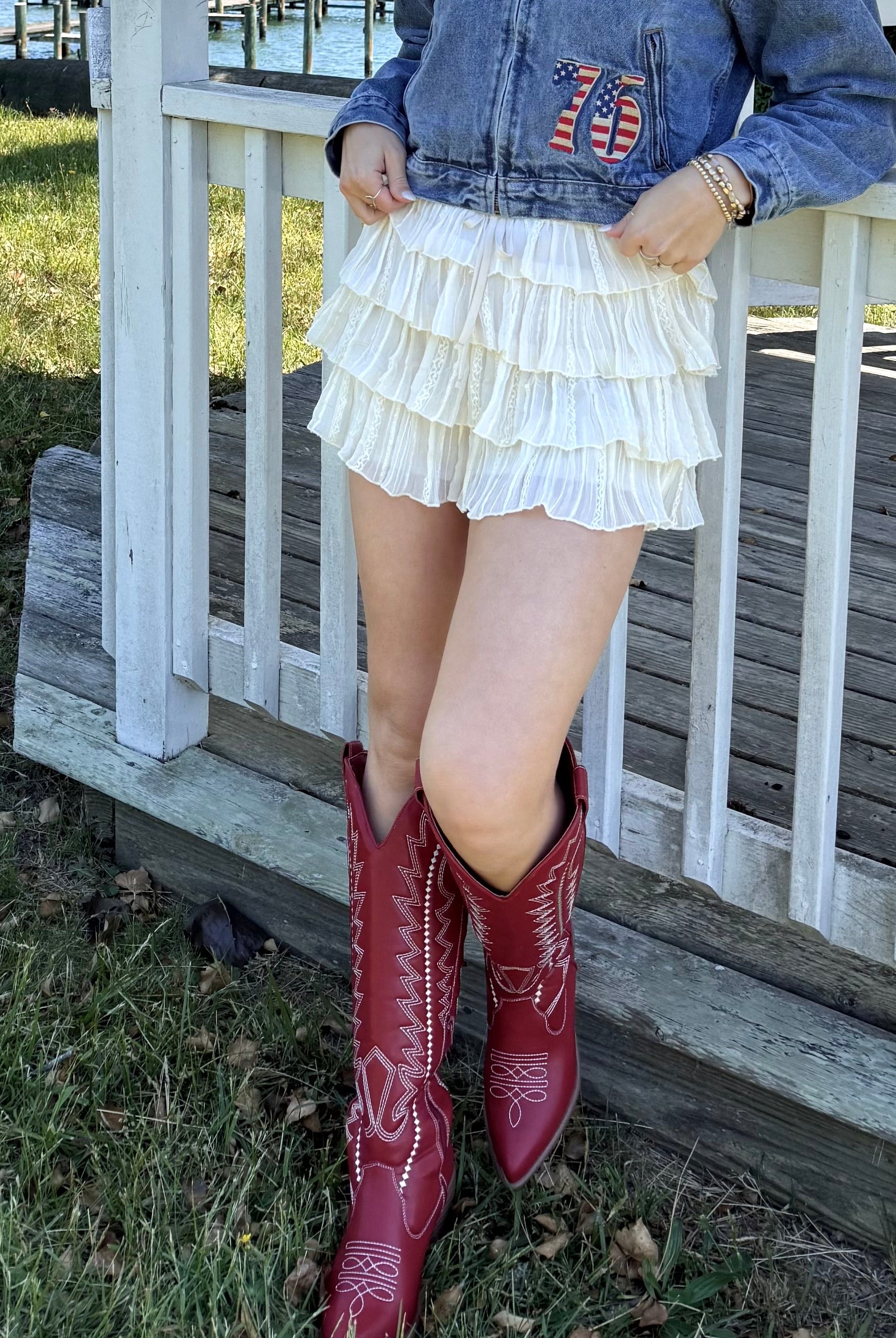 Woman in denim jacket and red boots standing on a porch by a lake, cream, airy, flowy, breathable, comfy, effortless, shimmer skirt, mini