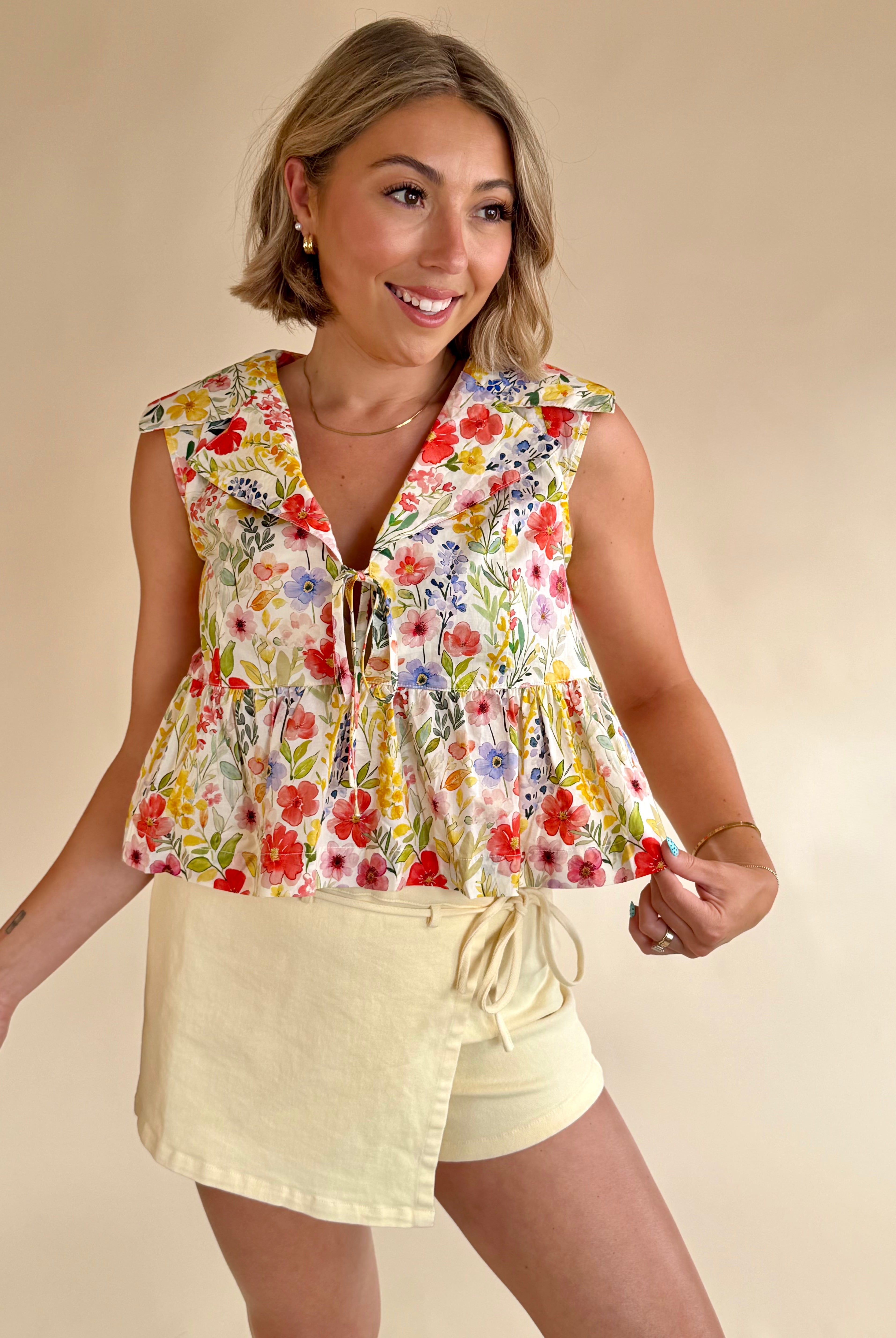 Woman wearing a floral sleeveless top and beige shorts on a beige background, flower pattern top, staple piece, rainbow shades of flowers, colorful tank, classy and girly