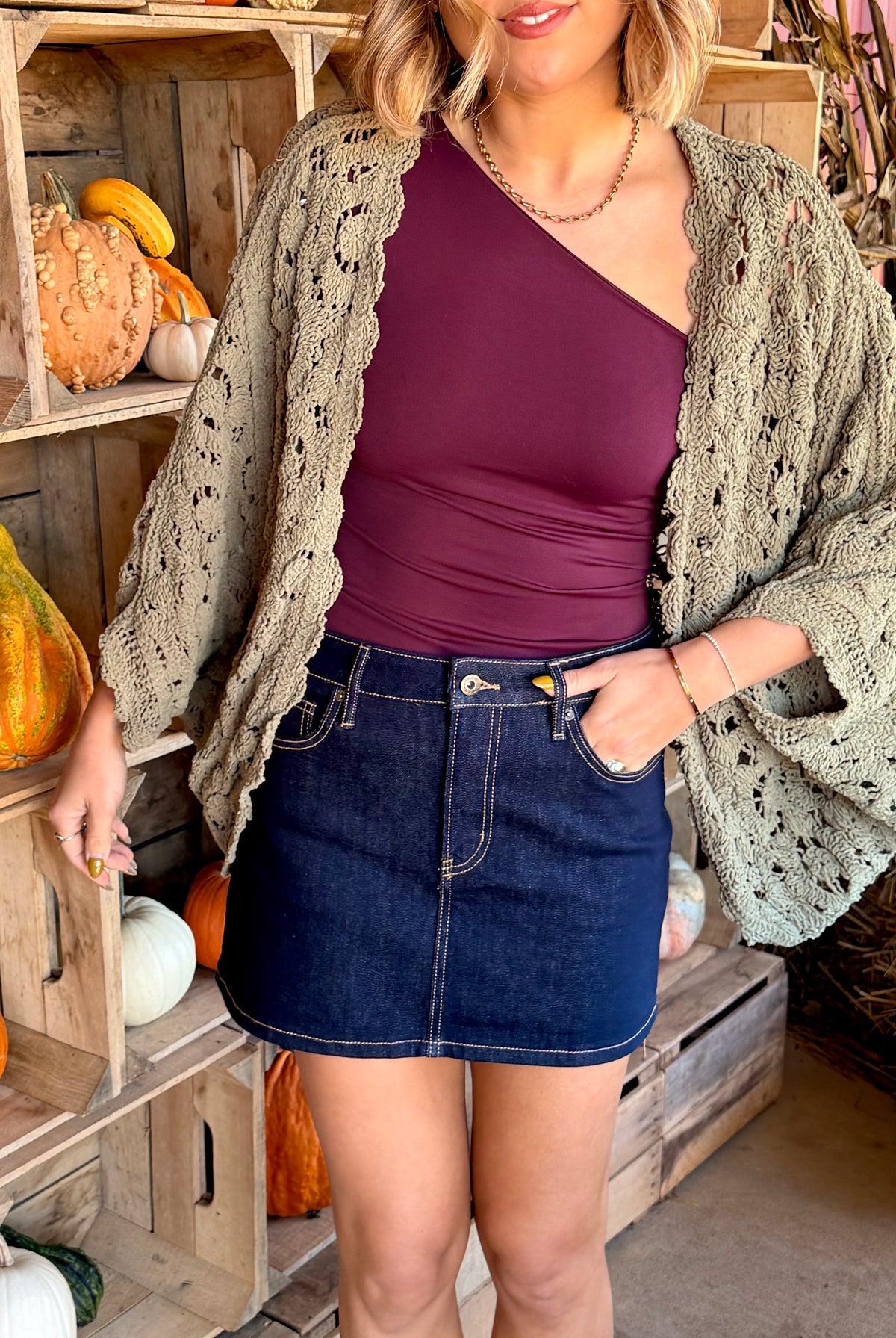 Model wearing an olive green chenille crochet cardigan with an openwork floral pattern, open front, scalloped edges, and batwing sleeves, layered over a plum top and dark denim mini skirt.