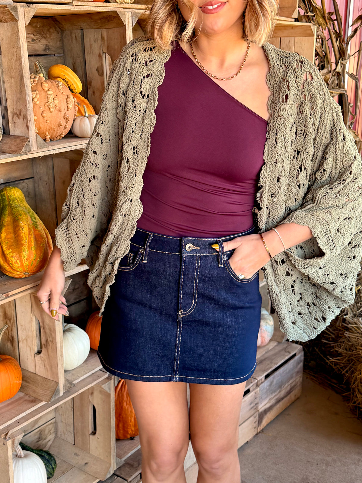 Model wearing an olive green chenille crochet cardigan with an openwork floral pattern, open front, scalloped edges, and batwing sleeves, layered over a plum top and dark denim mini skirt.
