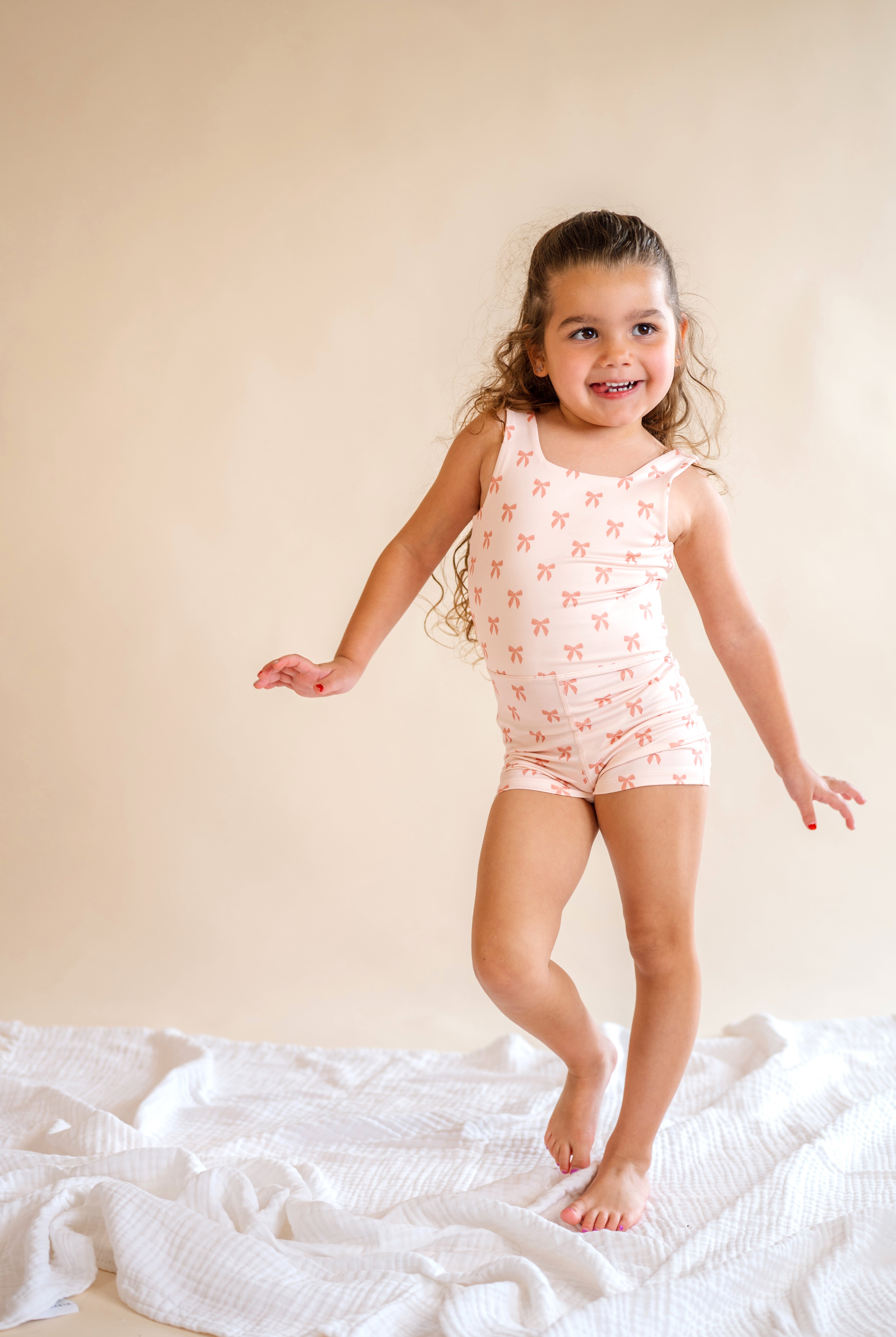 Young girl in a pink patterned onesie standing on a white blanket against a beige wall, pink bow body suit, one piece, kids clothes, pink bows, girly, kids athletic wear, summer wear