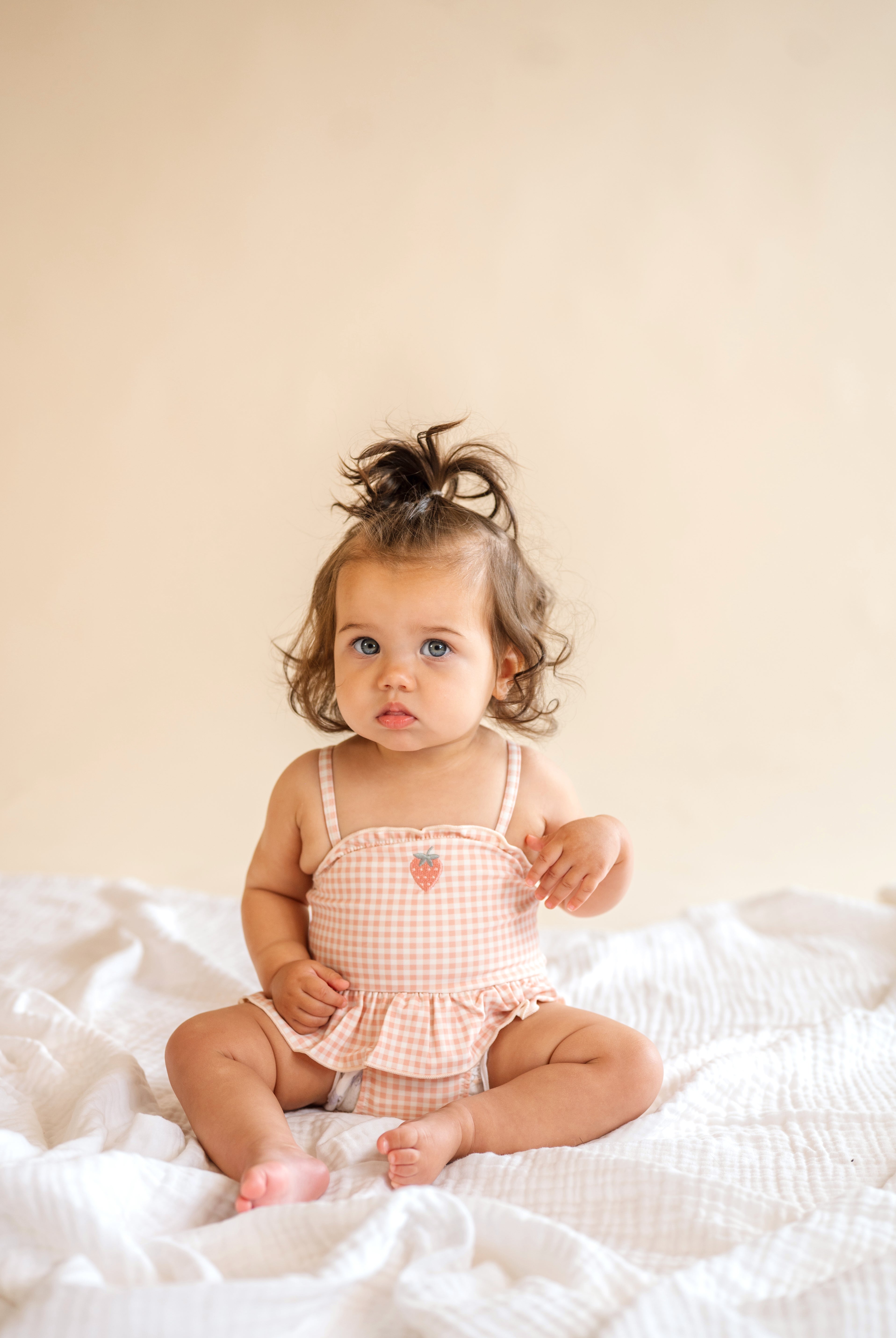 Baby sitting on a white blanket wearing a checkered romper against a beige background, strawberry patch one piece, baby swimwear, ruffle and spaghetti strap, pink plaid bathing suit