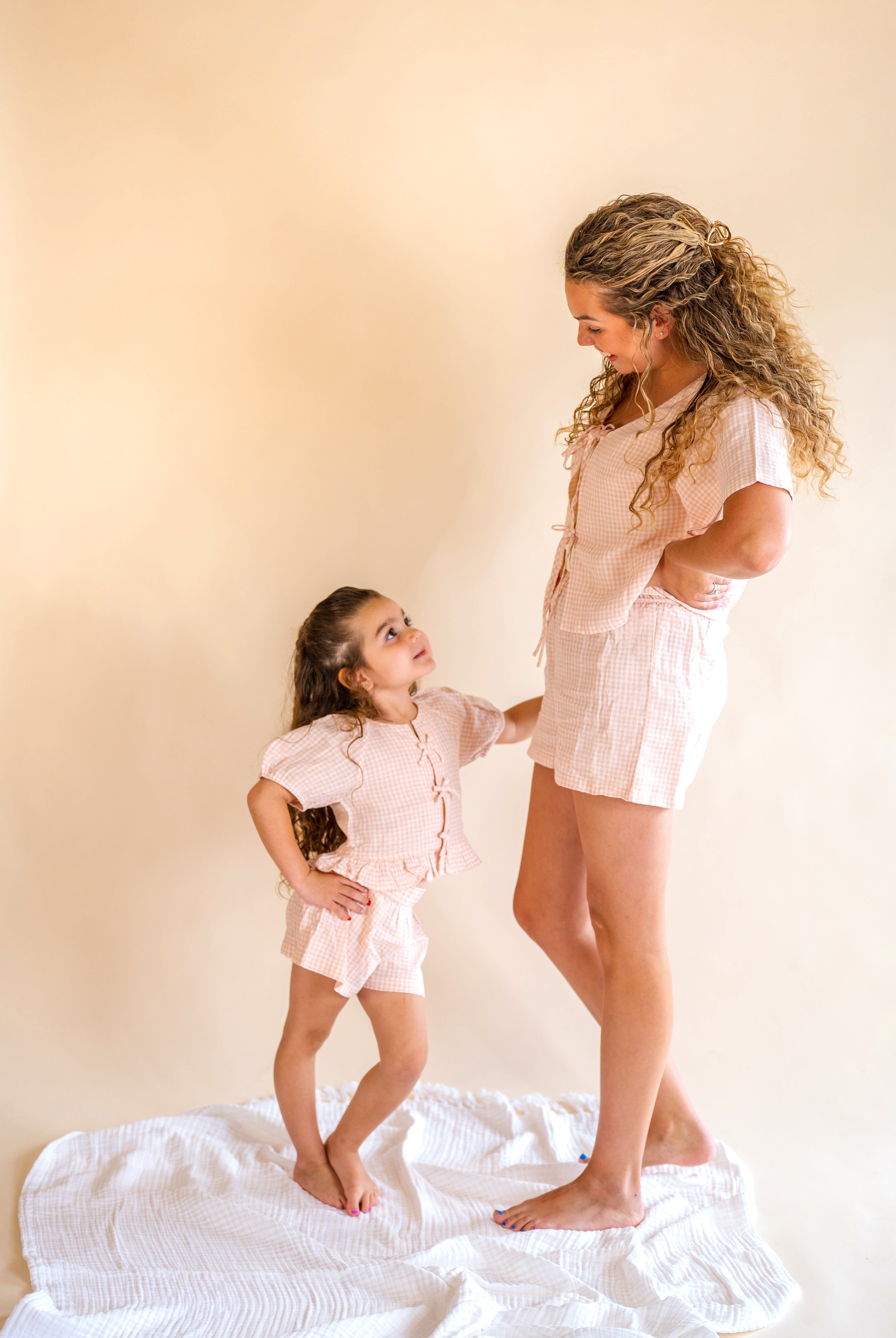 Woman and young girl in matching light pink outfits standing on a white surface with a beige background, mommy and me, pink plaid set, tie front top, plaid short set, kids wear