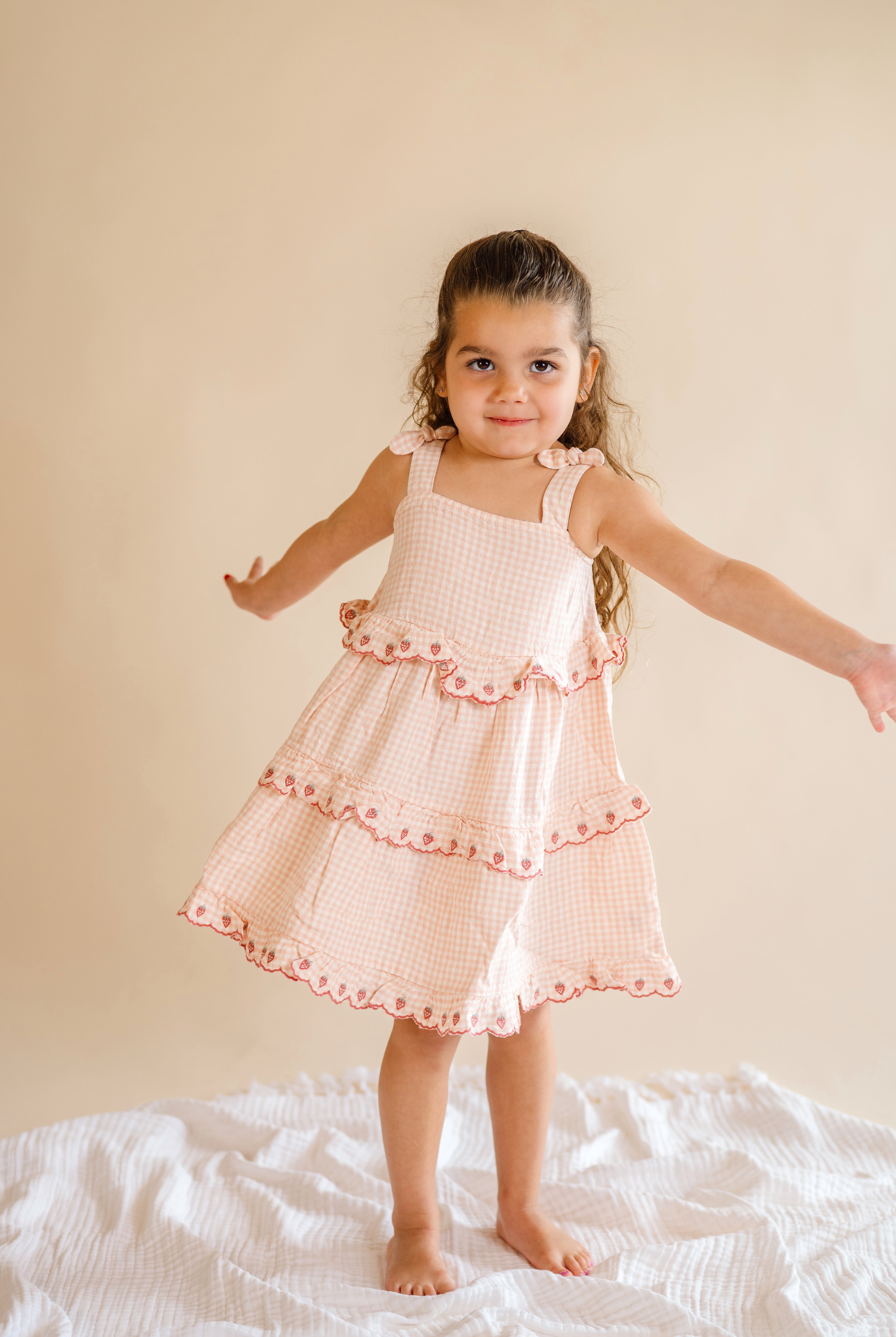Young girl in a pink checkered dress standing on a white surface with a beige background, strawberry ruffle dress, pink and plaid dress, ruffle tiered kid dress, pink and strawberry dream dress