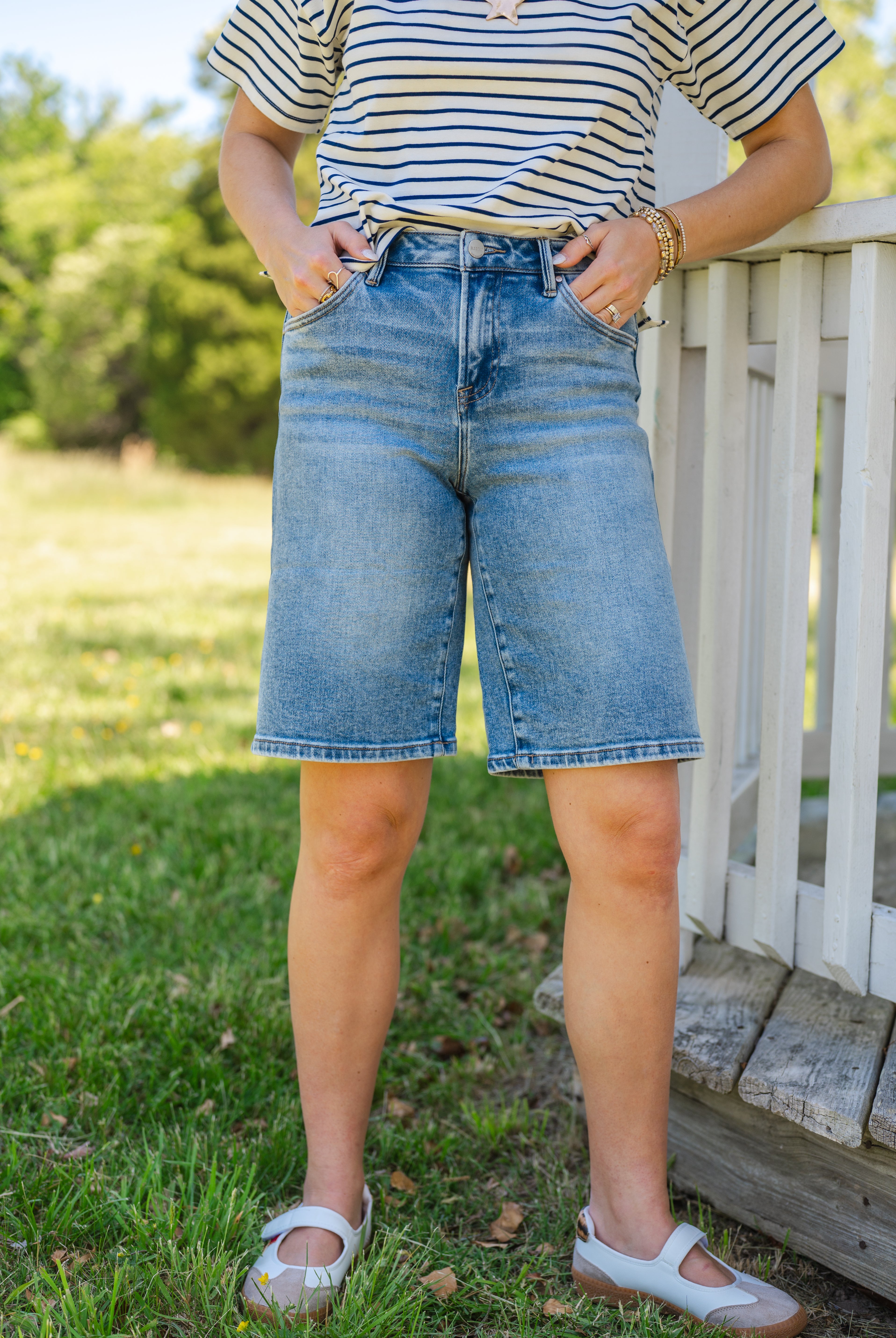 Person wearing a striped shirt and denim shorts standing on grass with a wooden fence in the background, Jort Style, long-length denim, universal summer short
