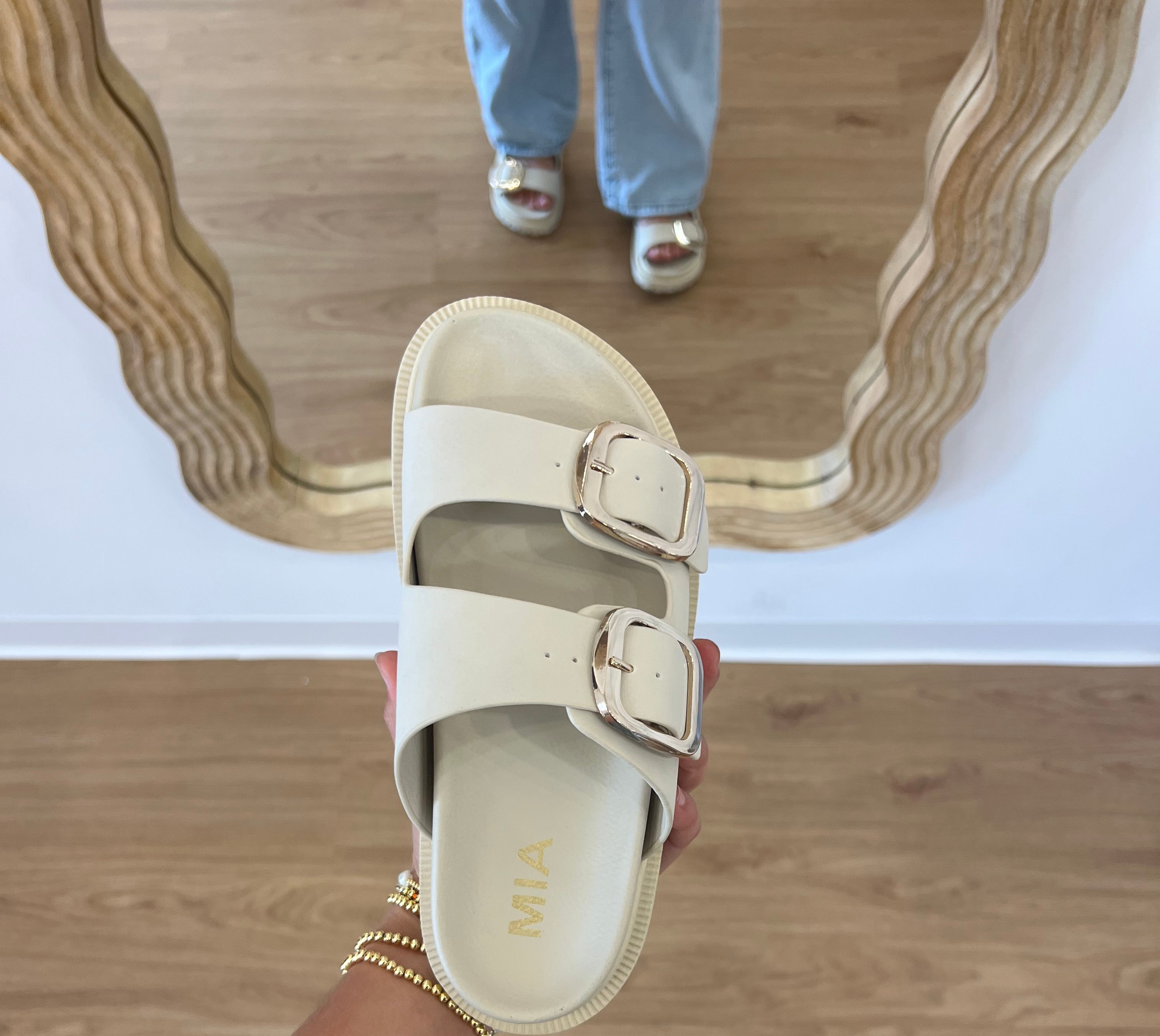 Person holding a beige sandal with 'Mia' branding on a wooden floor, slide snadal, pool slides, bach sandals, comfy footwear, birkenstock slides, gold buckel sandal