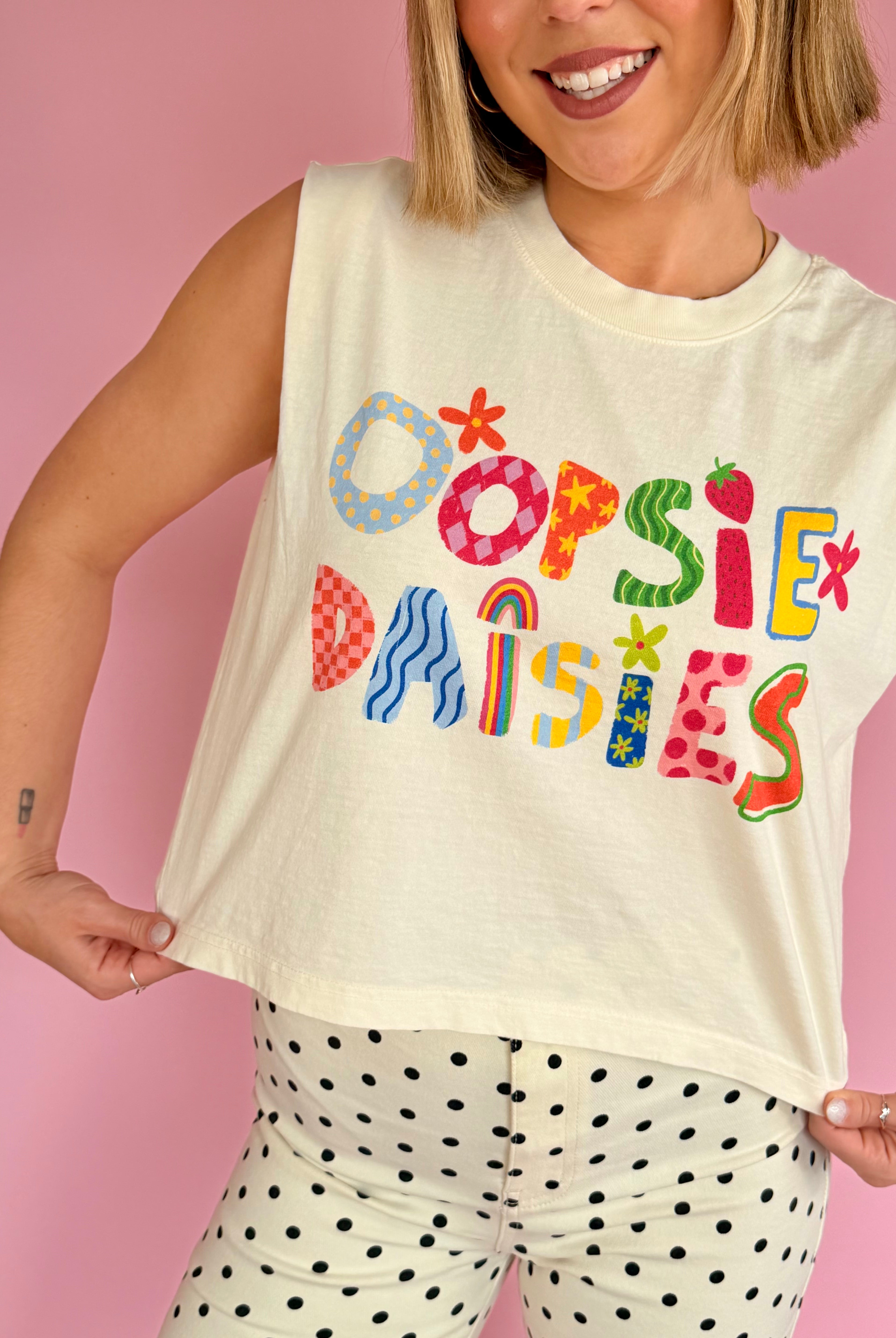 Person wearing a sleeveless shirt with colorful text 'OOPSIE DAISIES' against a pink background, pattern lover, sleeveless tee, flower and pattern contrast, cropped and flowy fit