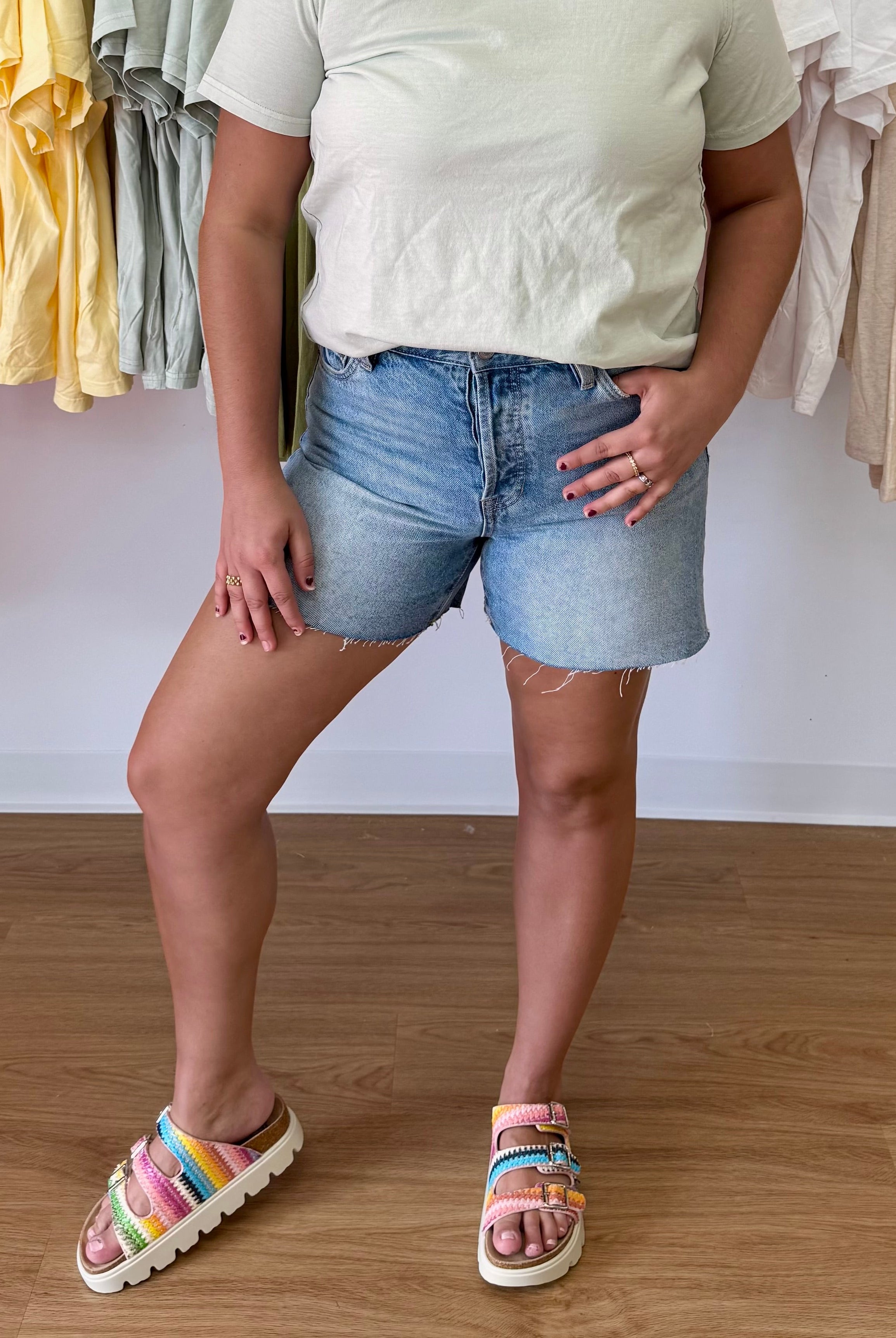 Person wearing a light-colored shirt and denim shorts standing on a wooden floor with clothing racks in the background, raw hem, short, button fly, stretchy