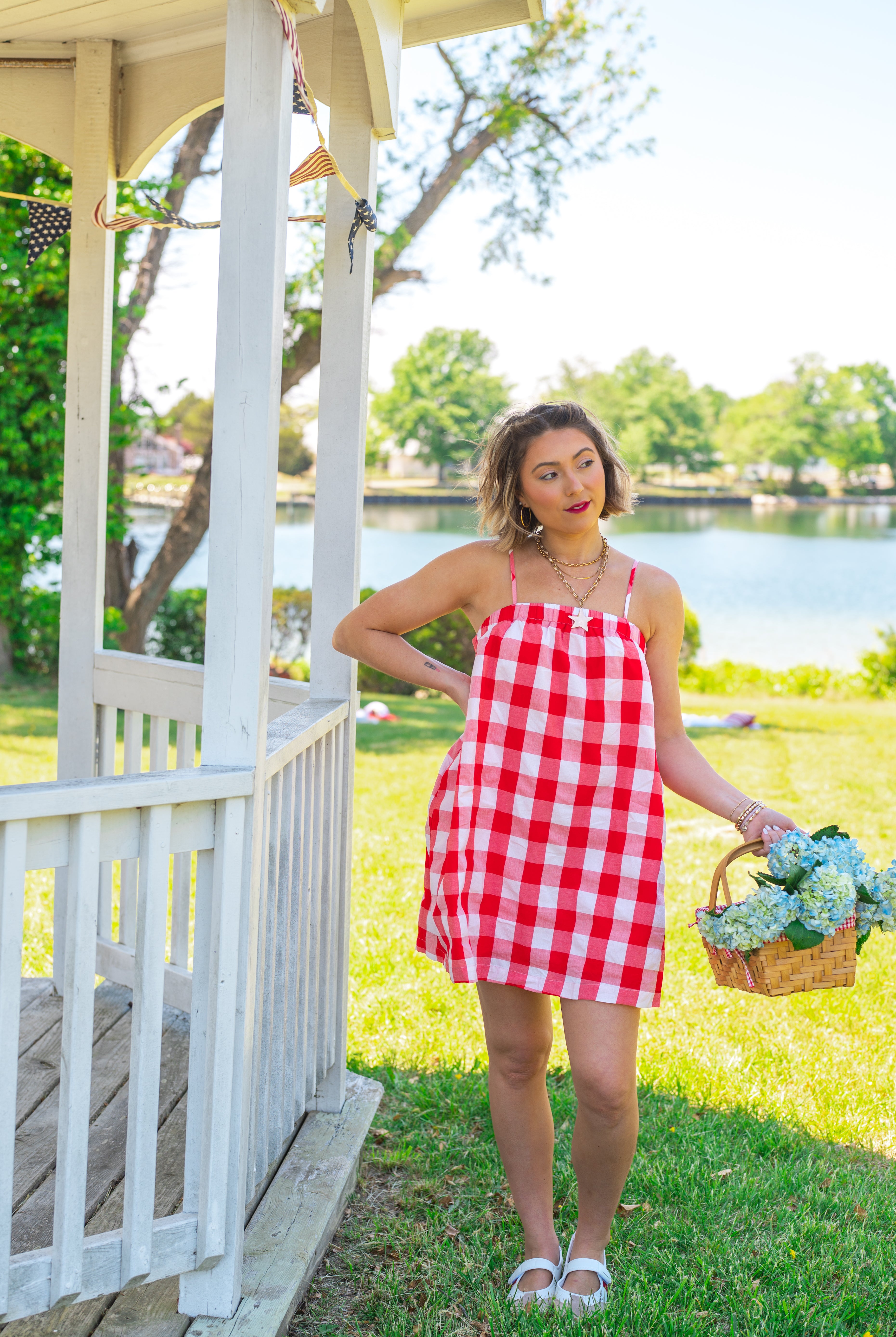 Woman in a red and white checkered dress standing by a gazebo with a lake in the background, red gingham, midi length, pockets, Americana, picnic cutie, flowy, straight fit,