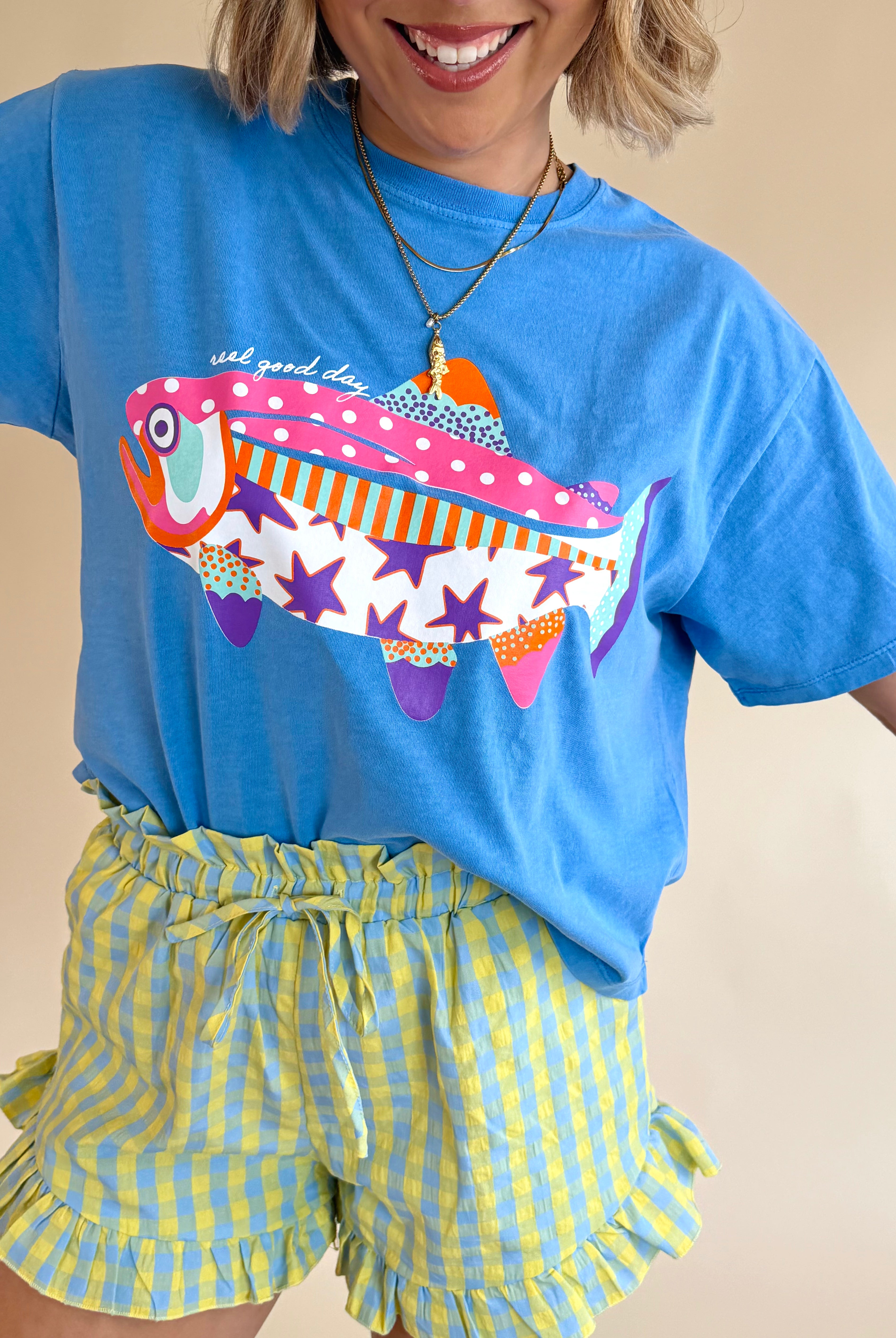 Person wearing a blue t-shirt with a colorful fish design and yellow checkered shorts on a beige background, patchwork tee, stars and polka dot, fish graphic tee, summer block color tee
