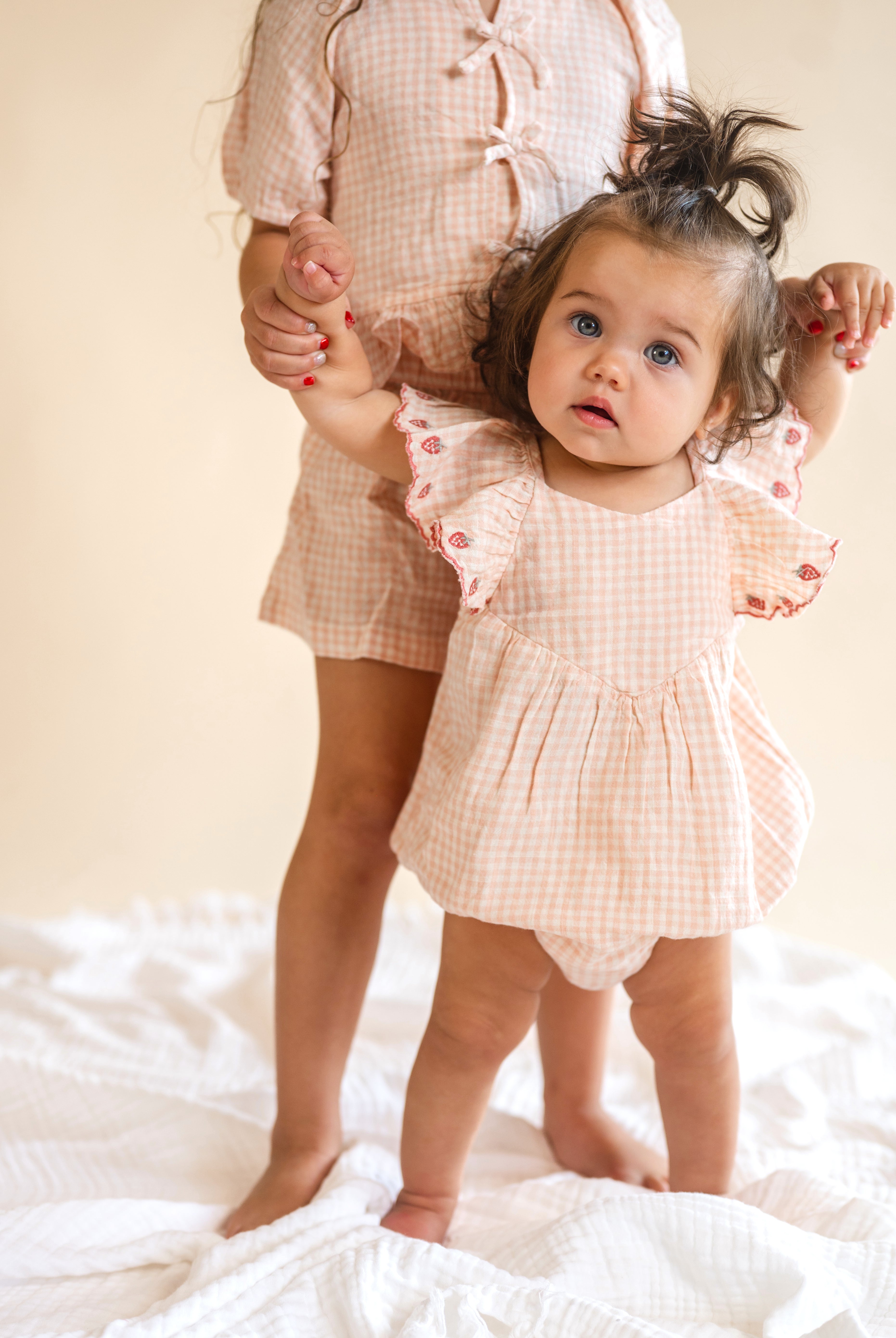 Child in a matching checkered outfit standing on a white surface with a neutral background, strawberry lined sleeves, flowy romper, baby wear, pink plaid