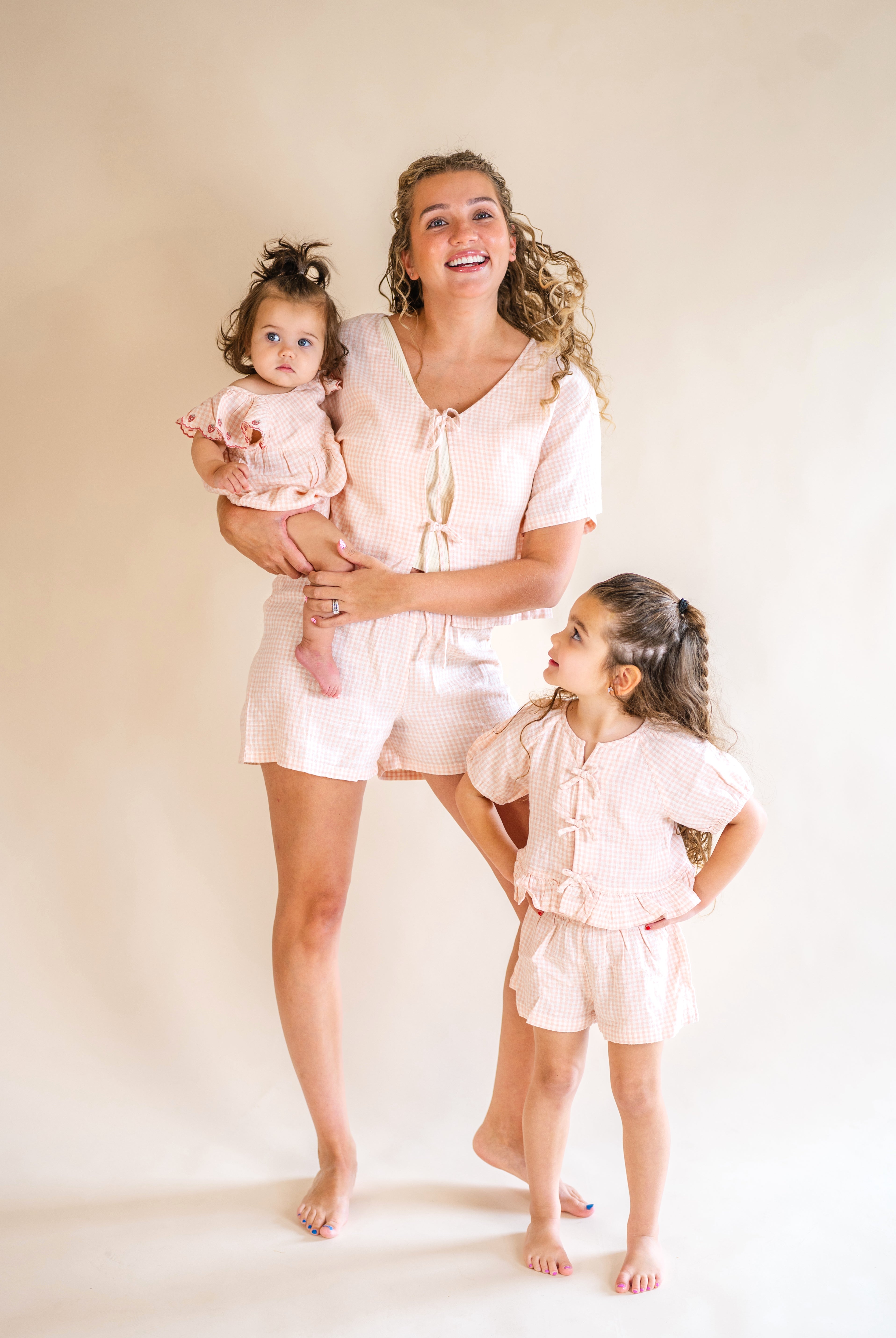 Woman and two young girls in matching light pink outfits standing against a plain background, pink plaid set, mommy and me, tie front top, plaid short set, pink and white matching set