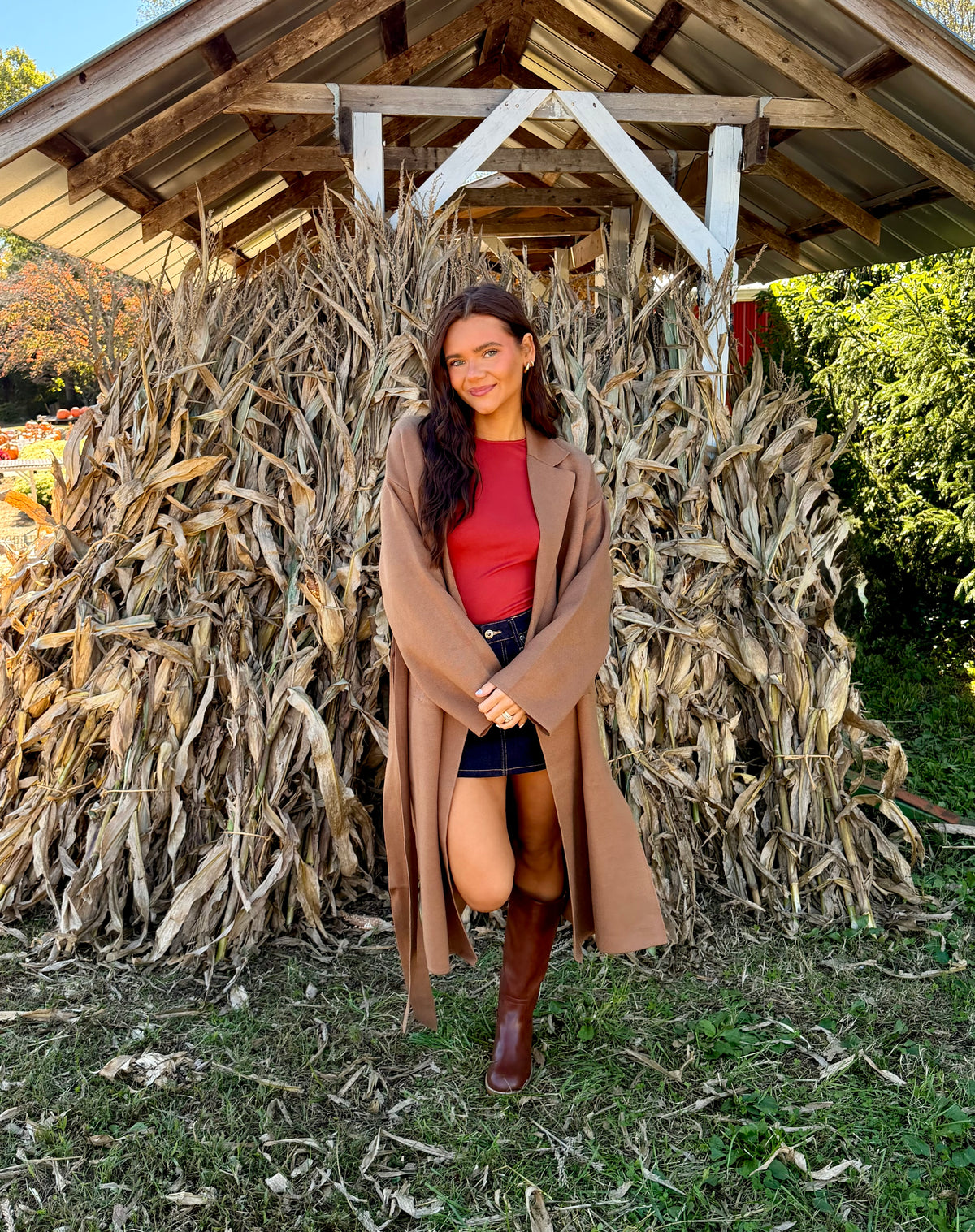 Model wearing a camel double-breasted wrap coat with a waist-cinching tie belt, long sleeves, and a trench-inspired longline silhouette.
