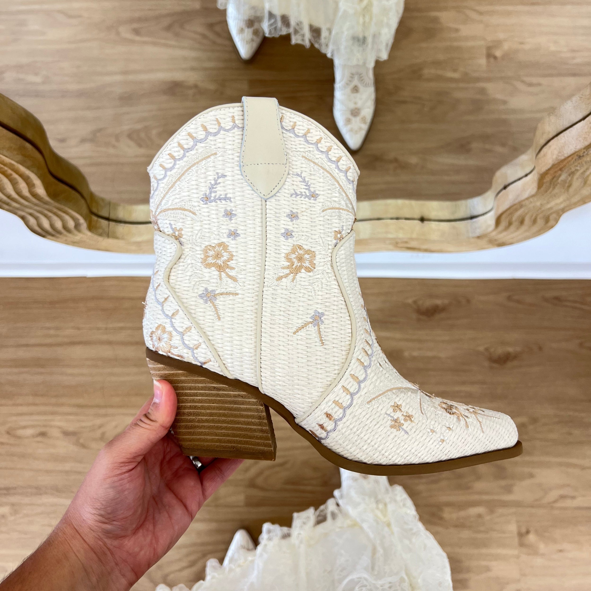 White embroidered boot held by a hand on a wooden floor with lace fabric. coastal cowgirl, raffia, western beach boot, COASTAL COWGIRL, WHITE, RAFFIA