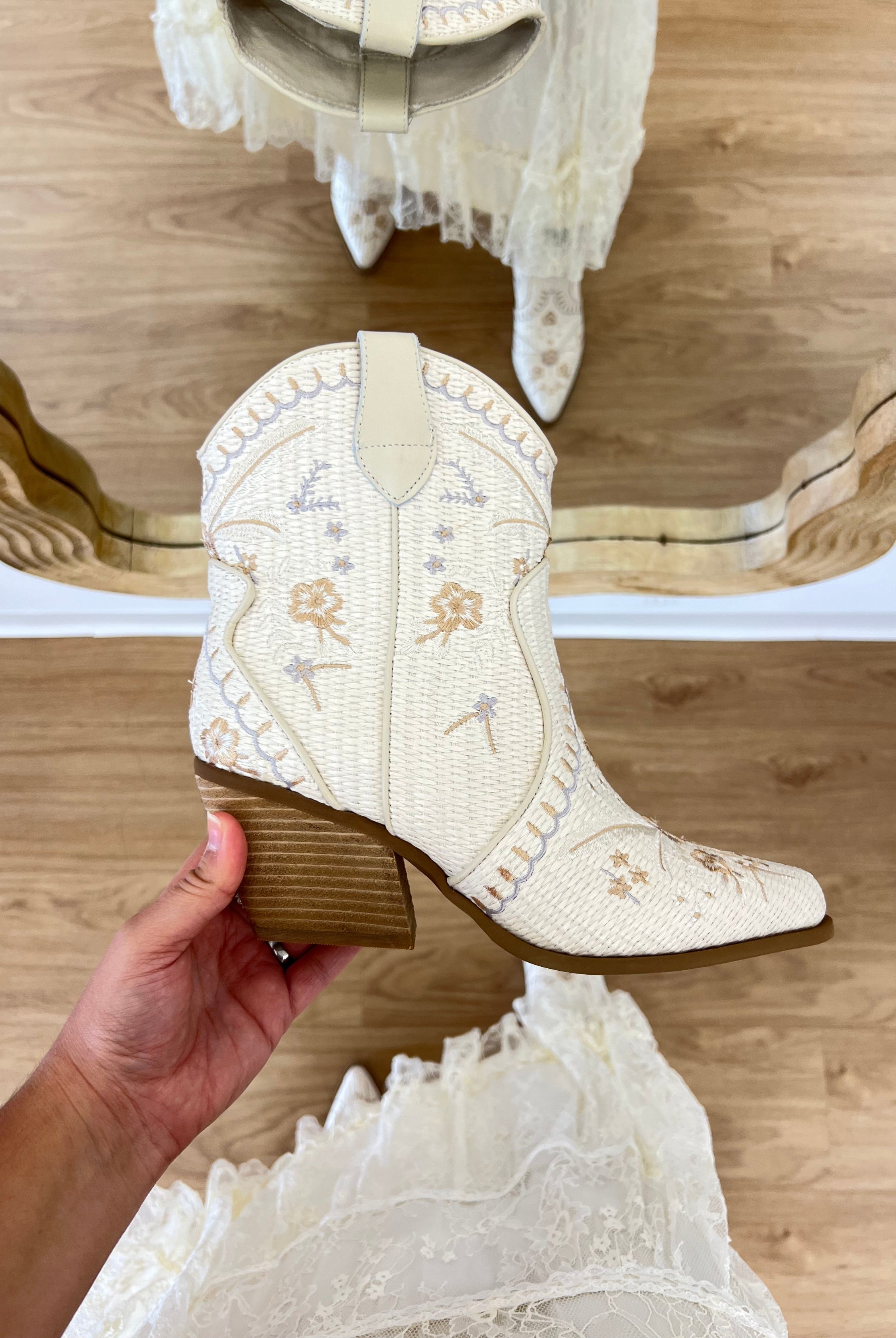 White embroidered boot held by a hand on a wooden floor with lace fabric. coastal cowgirl, raffia, western beach boot, COASTAL COWGIRL, WHITE, RAFFIA
