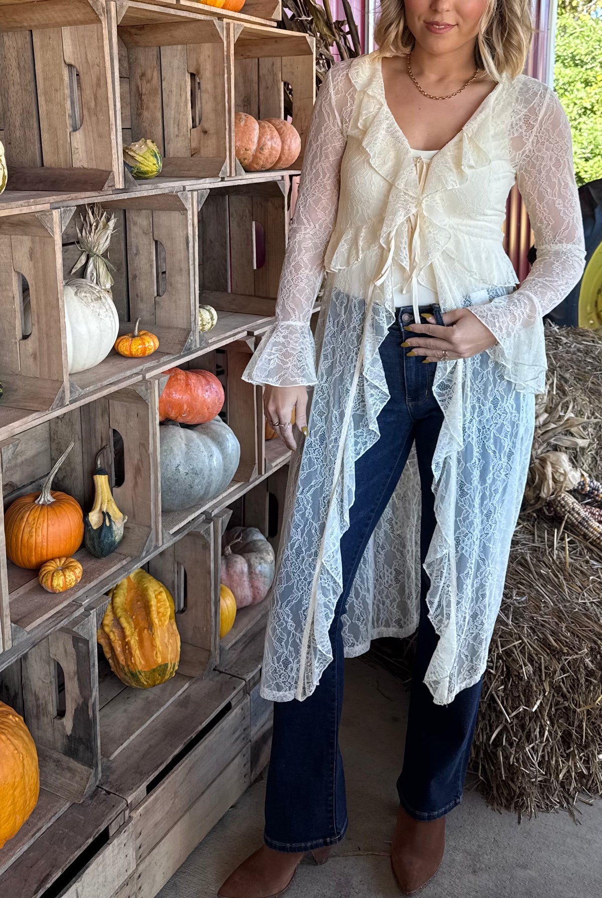Model wearing an ecru sheer lace duster with cascading ruffle trim, a tie-front closure, and long bell sleeves, layered over a white top and dark flare jeans.