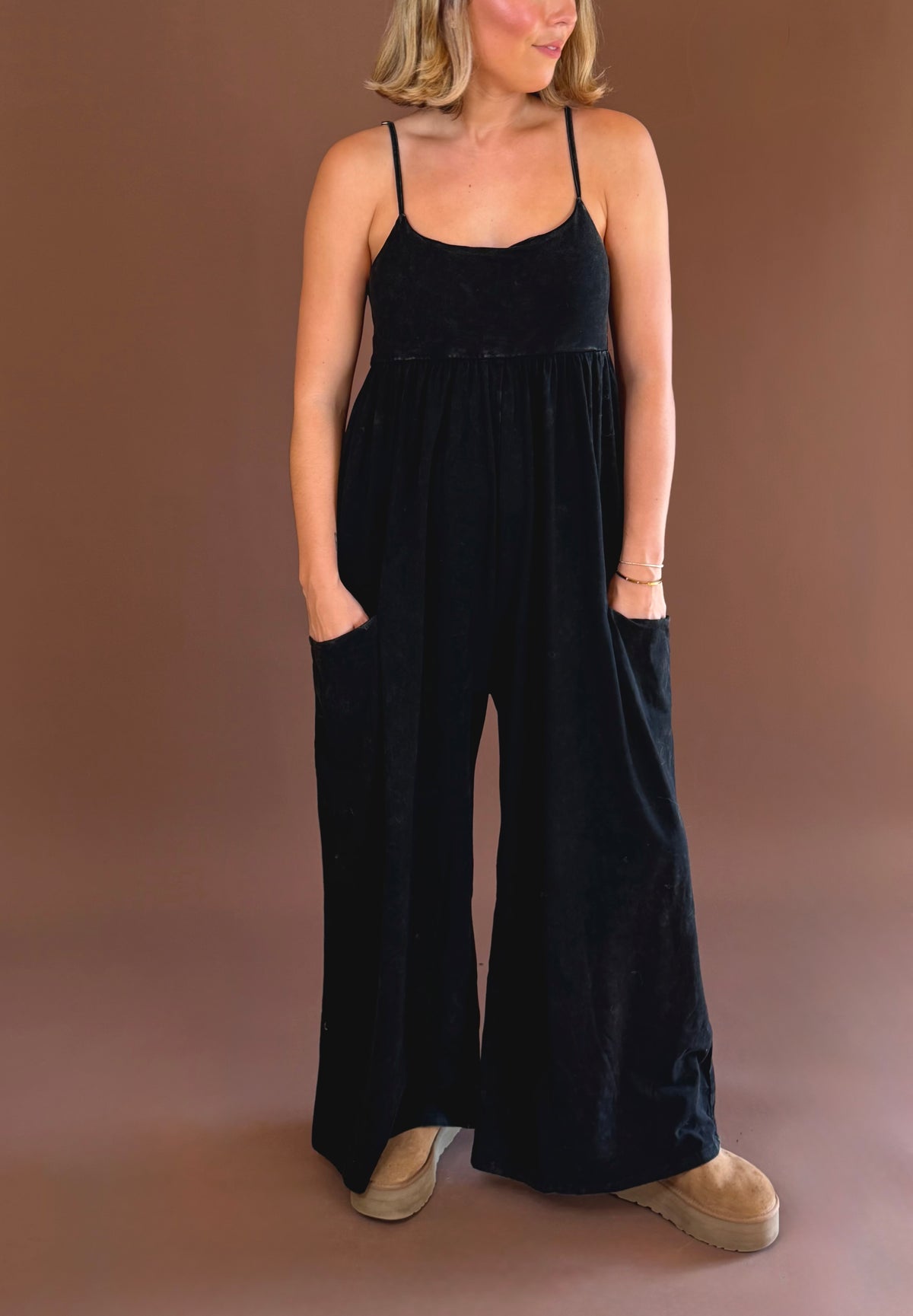 Model wearing a black wide leg jumpsuit with thin shoulder straps and front pockets, styled with brown clogs.