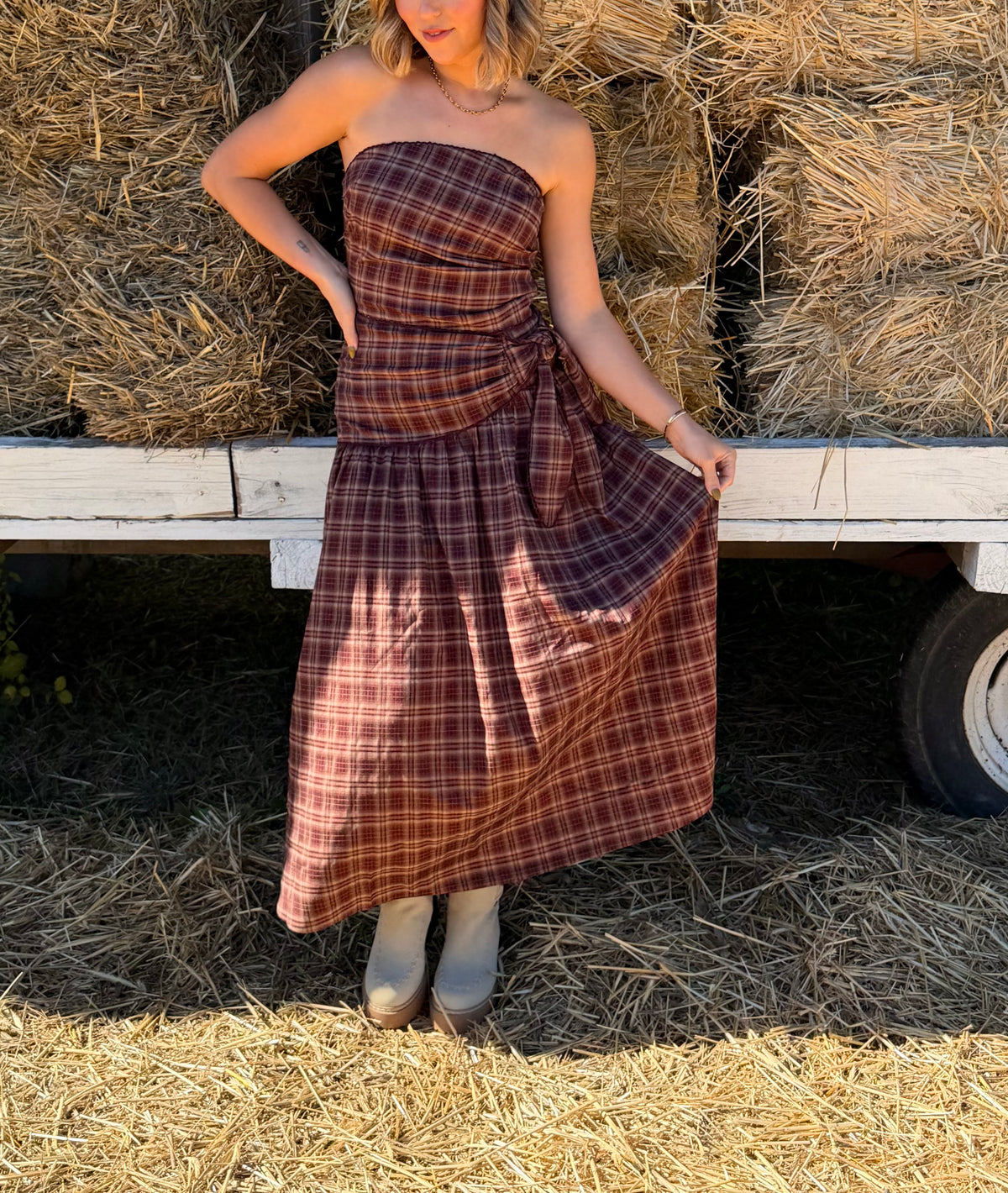 Model wearing a burgundy multicolor plaid strapless wrap dress with an adjustable side tie and a flowy full-length skirt.