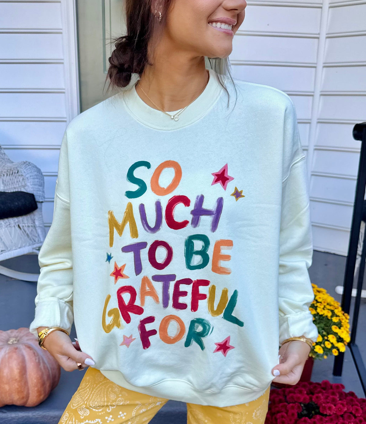 Model wearing a cream crewneck sweatshirt with colorful hand-lettered text that reads SO MUCH TO BE GRATEFUL FOR with star accents.