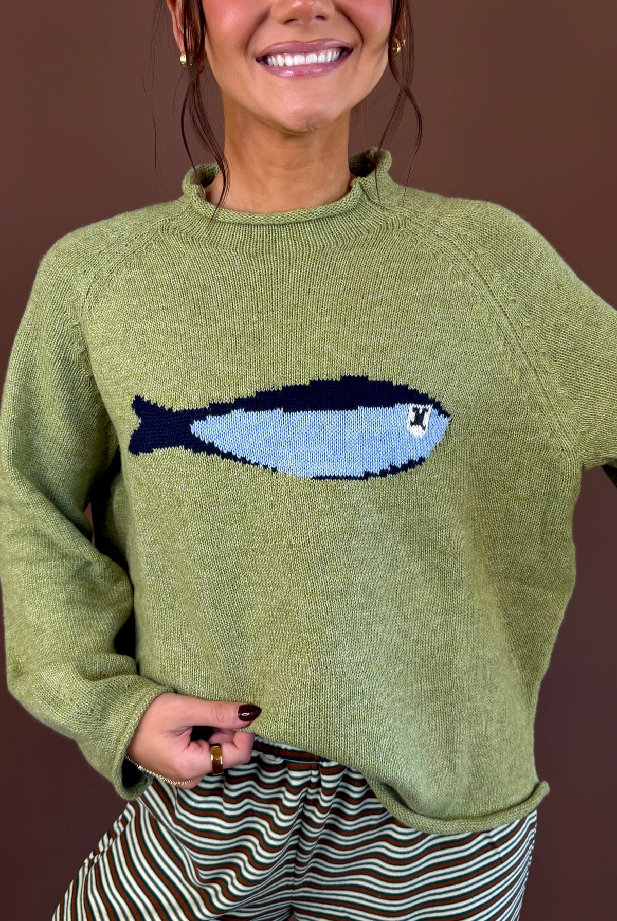 Model wearing an olive green mock neck sweater with a blue sardine graphic across the chest and long sleeves.