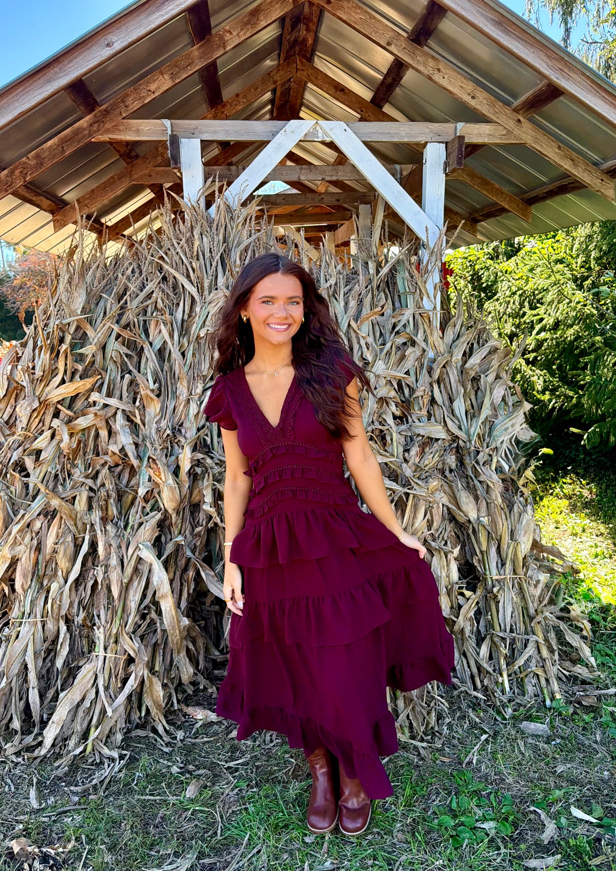 Model wearing a dark wine midi dress with a deep V-neckline, flutter short sleeves, a smocked waist, and a tiered ruffle skirt, styled with brown boots in front of dried cornstalks.