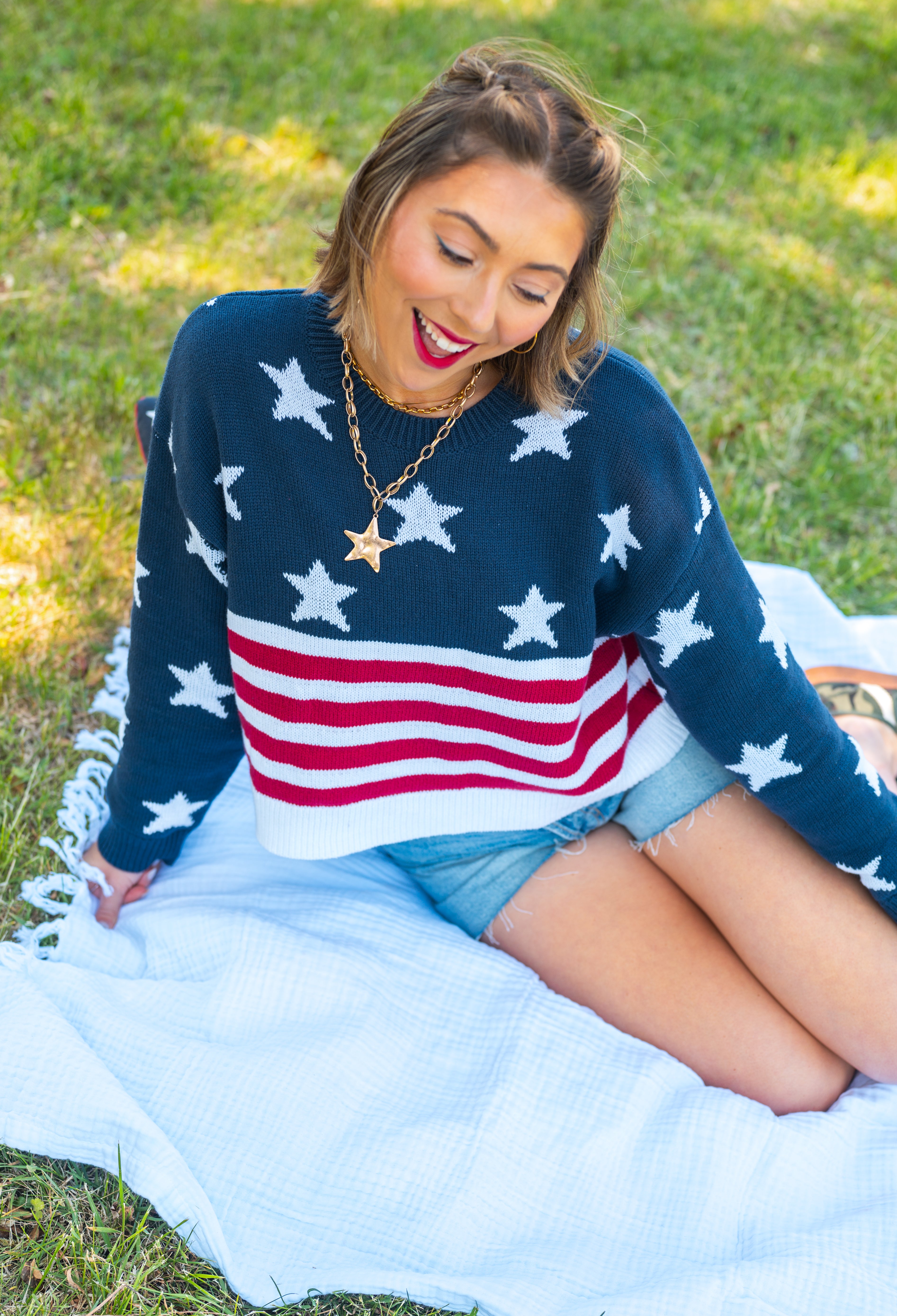 Woman wearing a sweater with an American flag design, sitting on grass, USA, cropped sweater, Americana, red white and blue, chunky sweater, stars and stripes