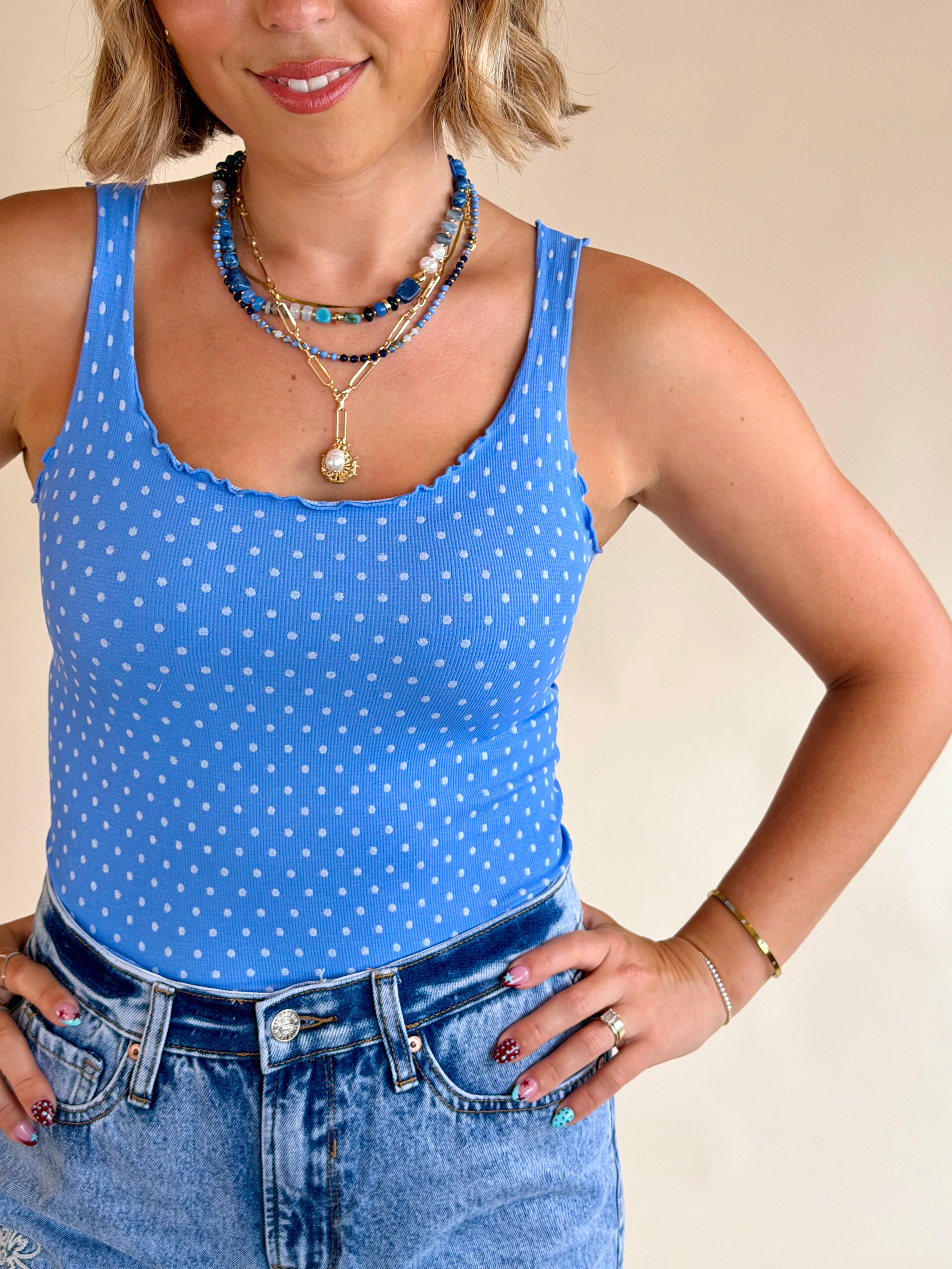 Woman wearing a blue polka dot tank top and jeans with a beige background, summer basic, ruffle hem tank, blue and white polka dot, nikibiki, flattering summer fit 