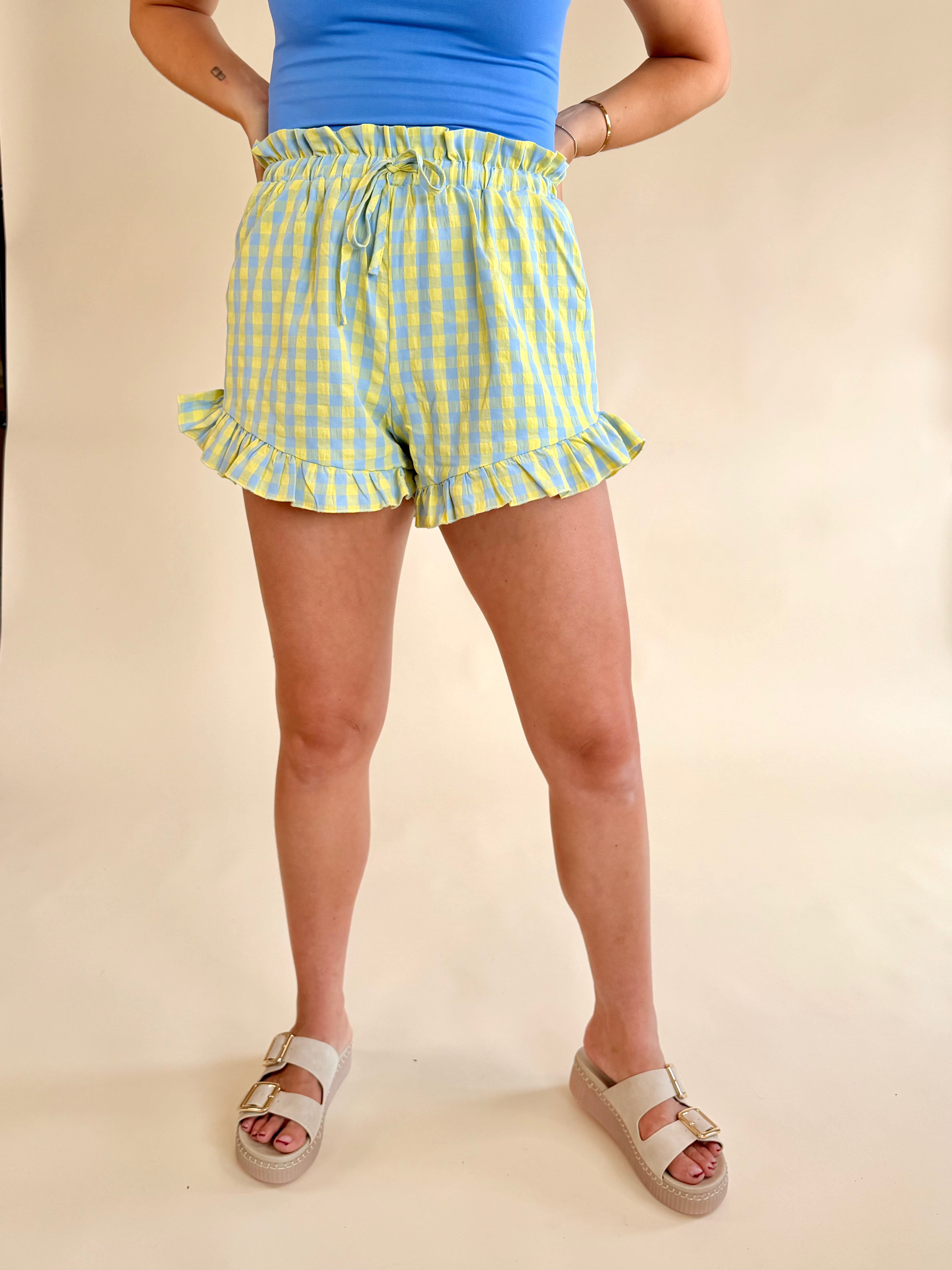 Person wearing yellow checkered shorts and a blue top on a beige background, Blue and yellow summer short, ruffle short, drawstring summer pants, pastels and neons, blue and green 