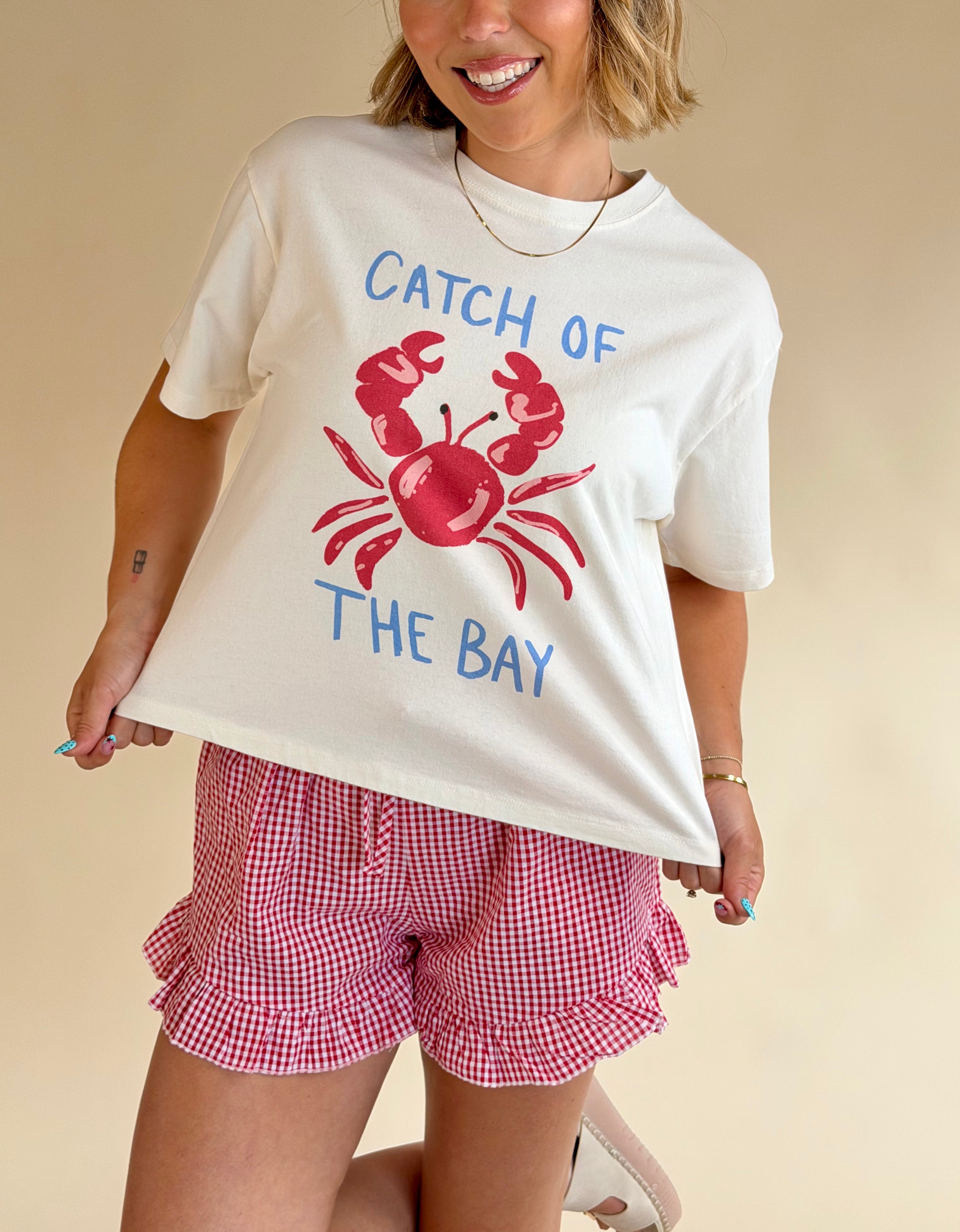 Person wearing a white t-shirt with a red crab graphic and text, paired with red checkered shorts on a beige background, crab graphic tee, cropped fit tee, red white and blue, summer staple, crab graphic 