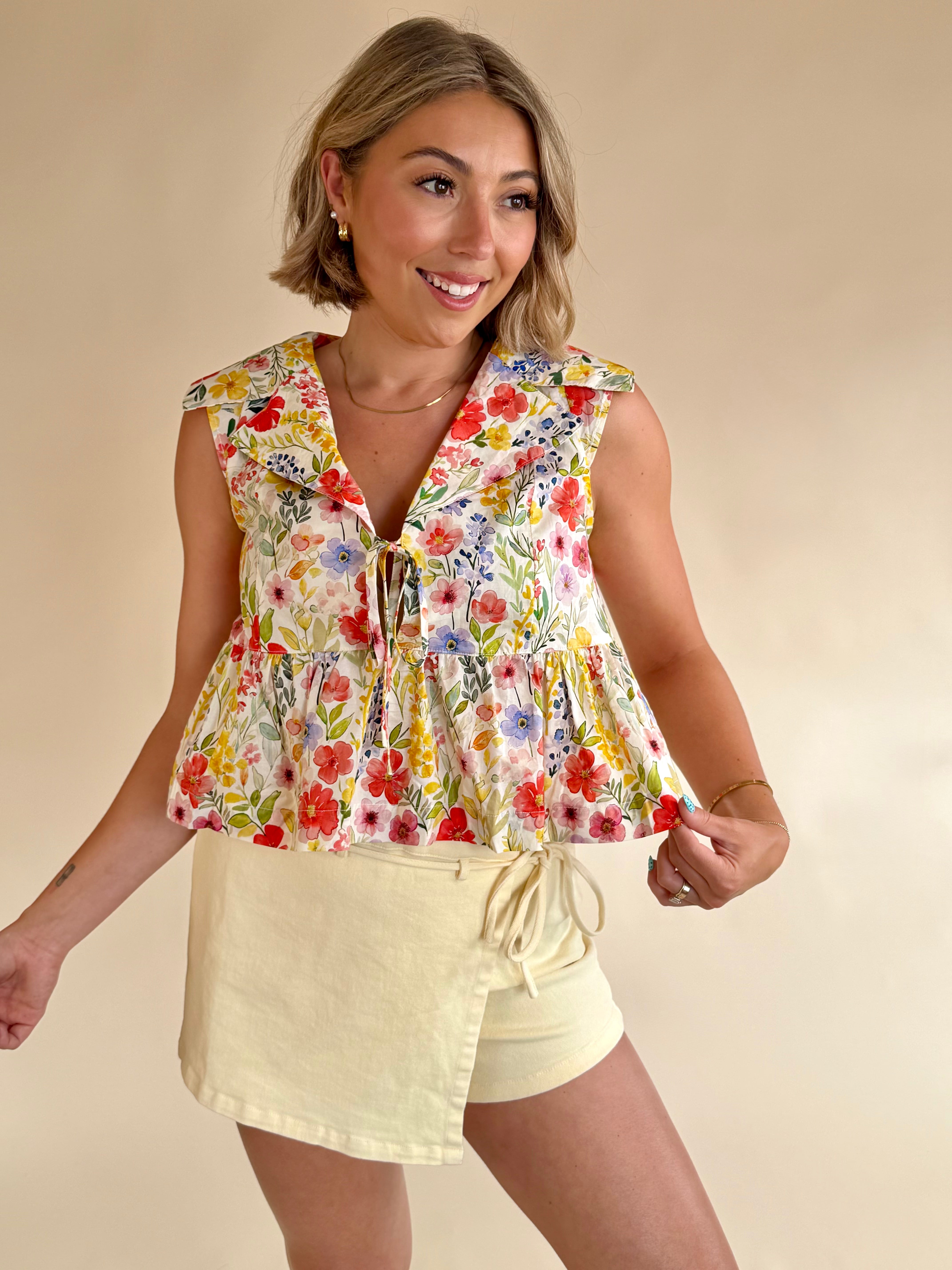 Woman wearing a floral sleeveless top and beige shorts on a beige background, flower pattern top, staple piece, rainbow shades of flowers, colorful tank, classy and girly 