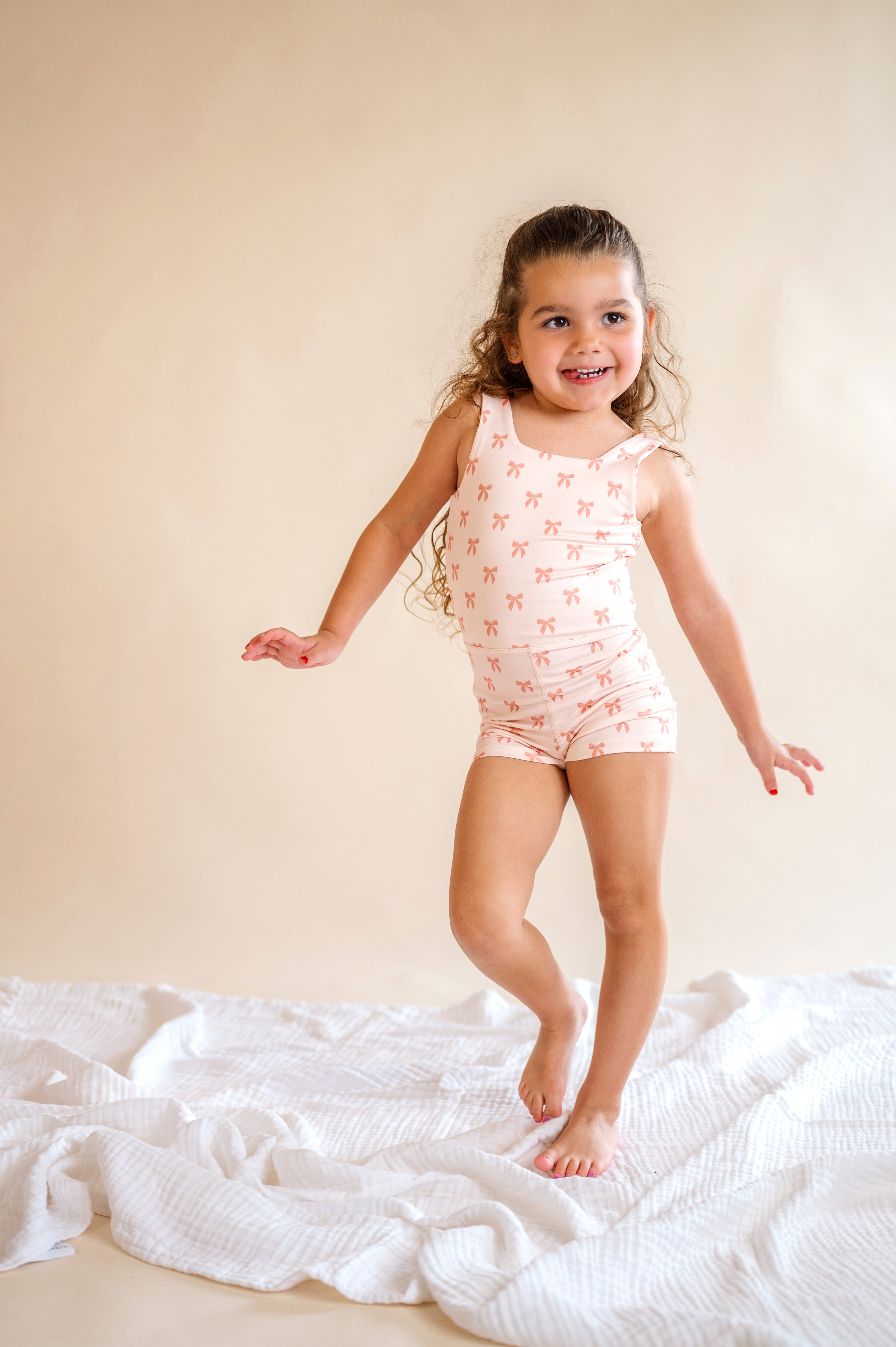 Young girl in a pink patterned onesie standing on a white blanket against a beige wall, pink bow body suit, one piece, kids clothes, pink bows, girly, kids athletic wear, summer wear