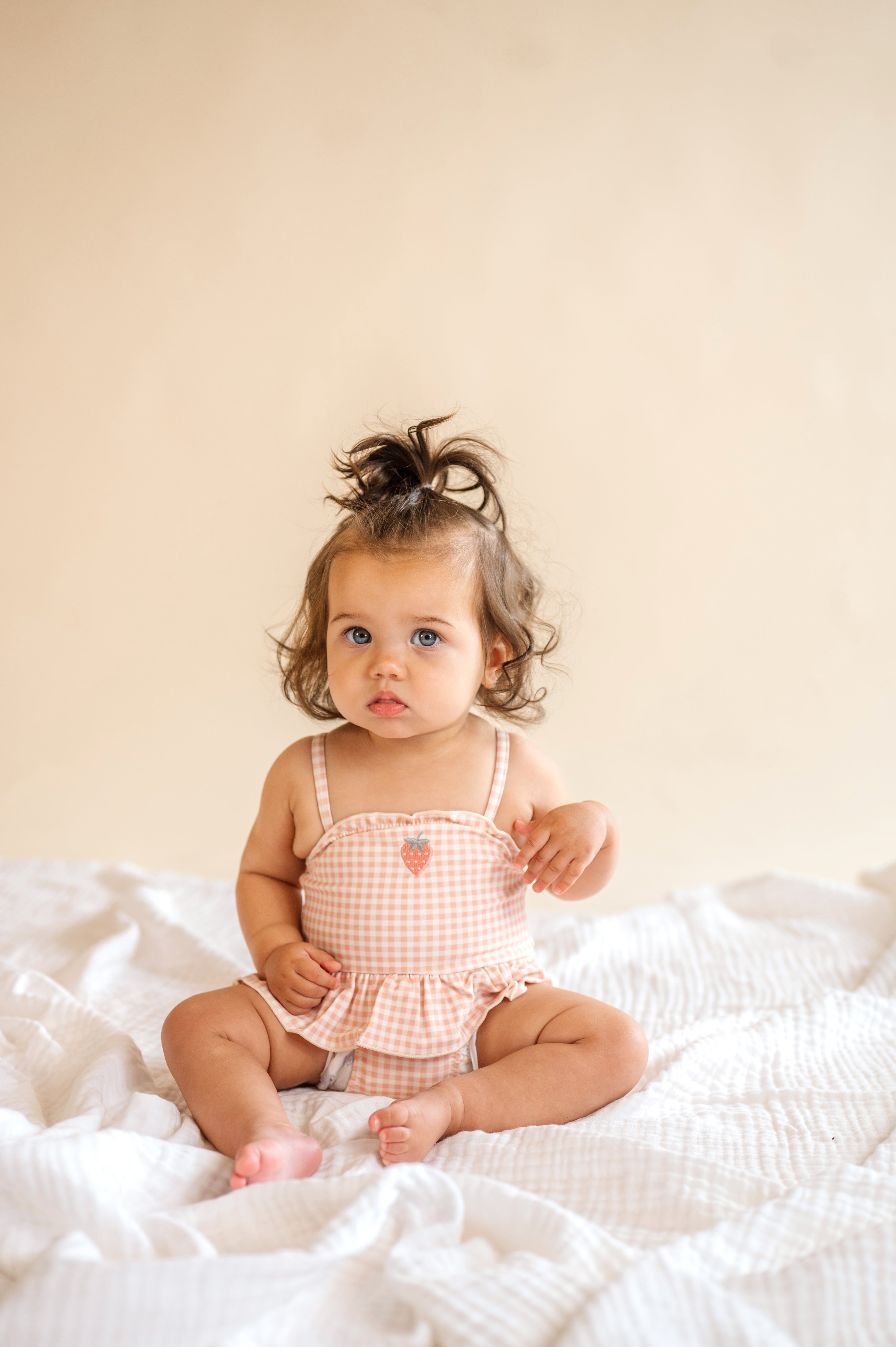 Baby sitting on a white blanket wearing a checkered romper against a beige background, strawberry patch one piece, baby swimwear, ruffle and spaghetti strap, pink plaid bathing suit 
