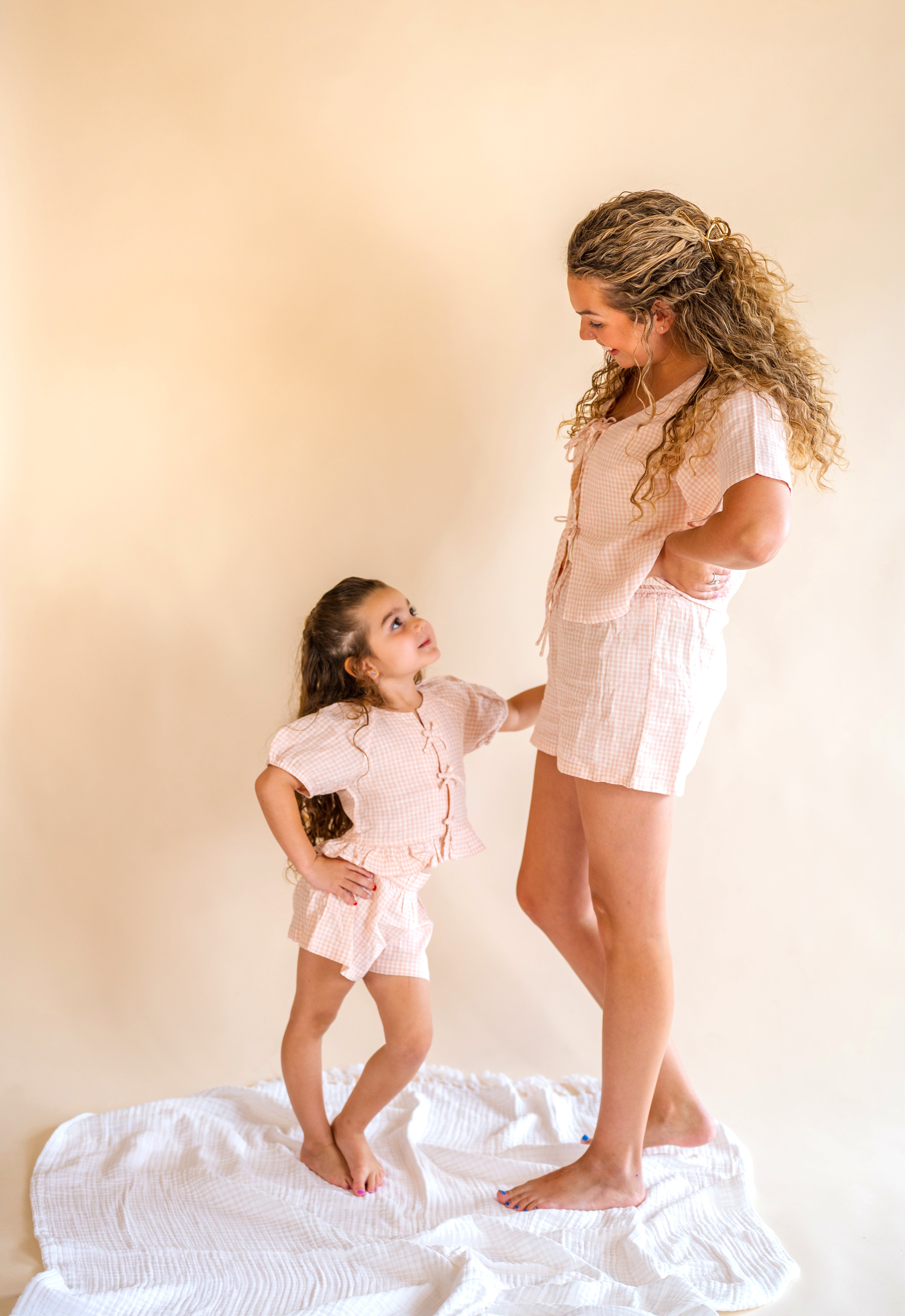 Woman and young girl in matching light pink outfits standing on a white surface with a beige background, mommy and me, pink plaid set, tie front top, plaid short set, kids wear
