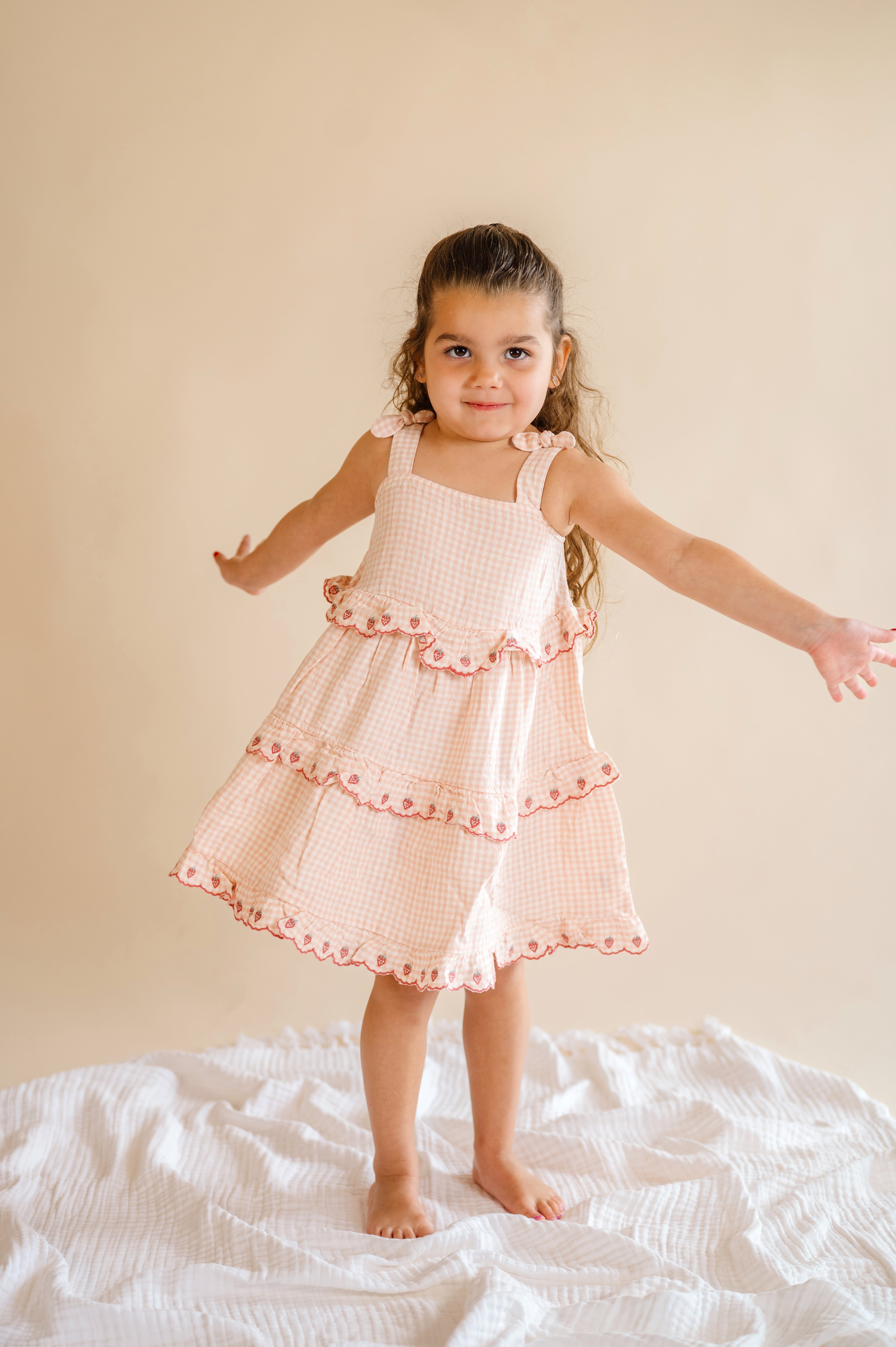 Young girl in a pink checkered dress standing on a white surface with a beige background, strawberry ruffle dress, pink and plaid dress, ruffle tiered kid dress, pink and strawberry dream dress 