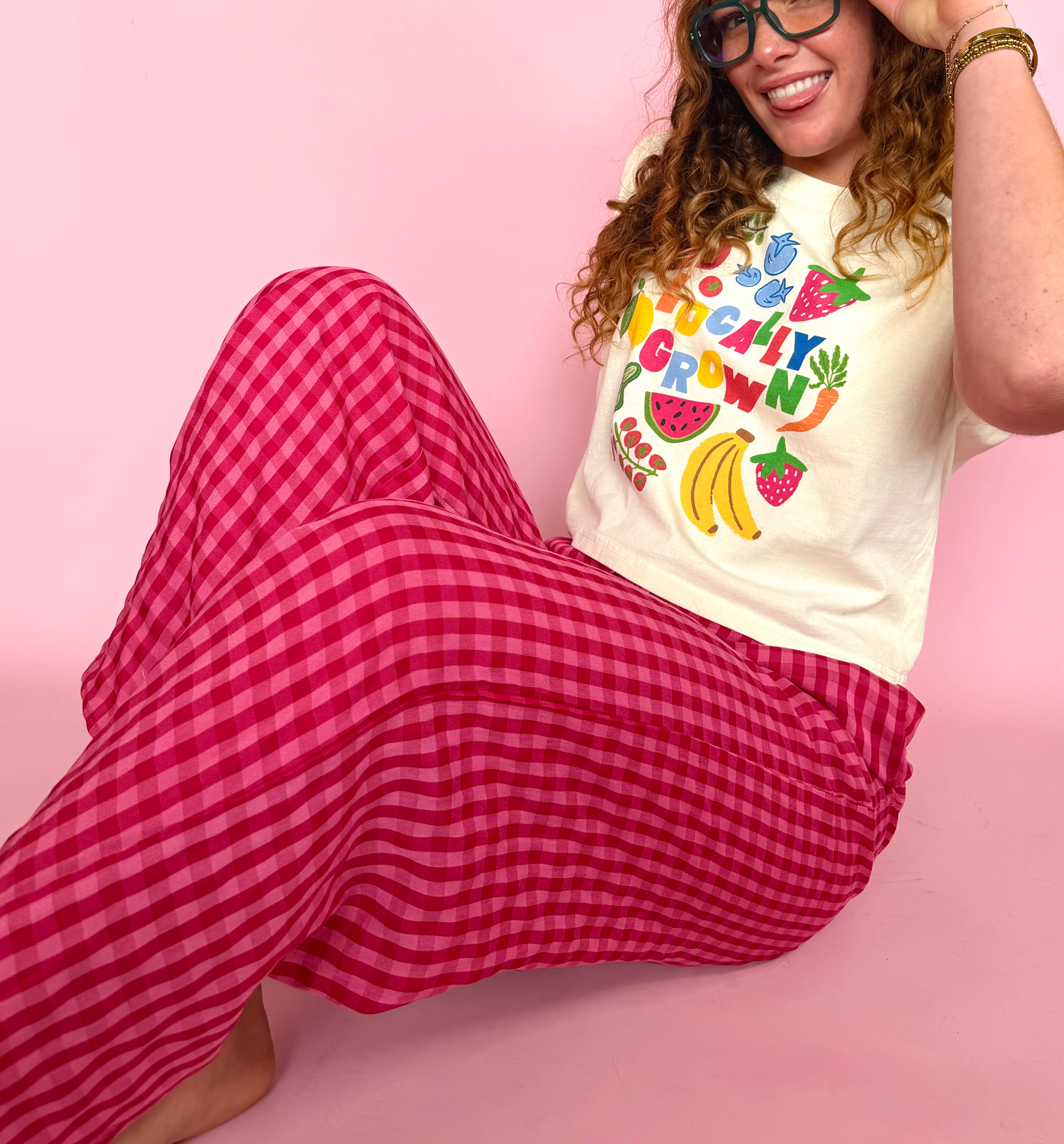 Person wearing a colorful t-shirt and red checkered pants on a pink background, fruits and veggies, colorful graphic tank, full length flowy fit