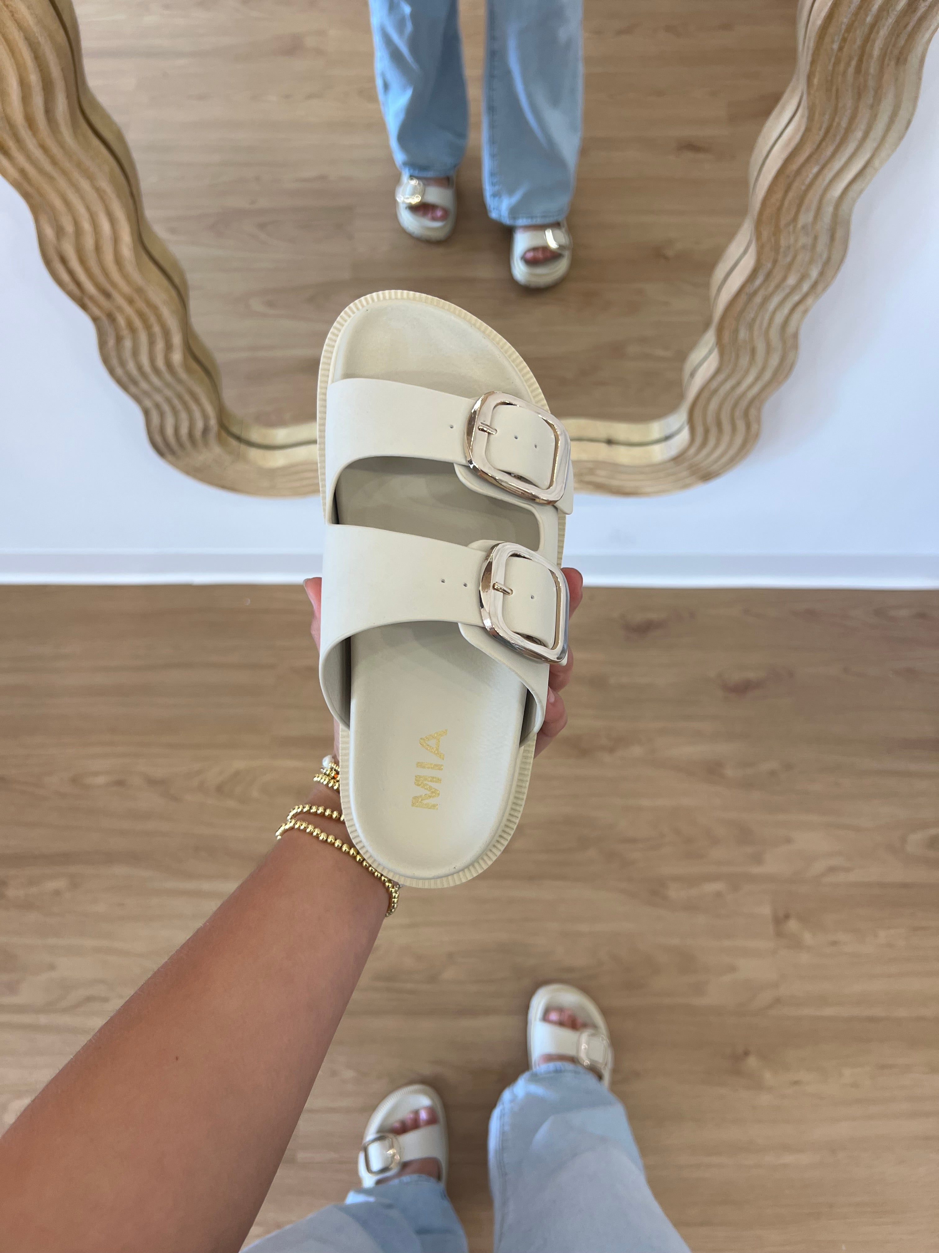 Person holding a beige sandal with 'Mia' branding on a wooden floor, slide snadal, pool slides, bach sandals, comfy footwear, birkenstock slides, gold buckel sandal