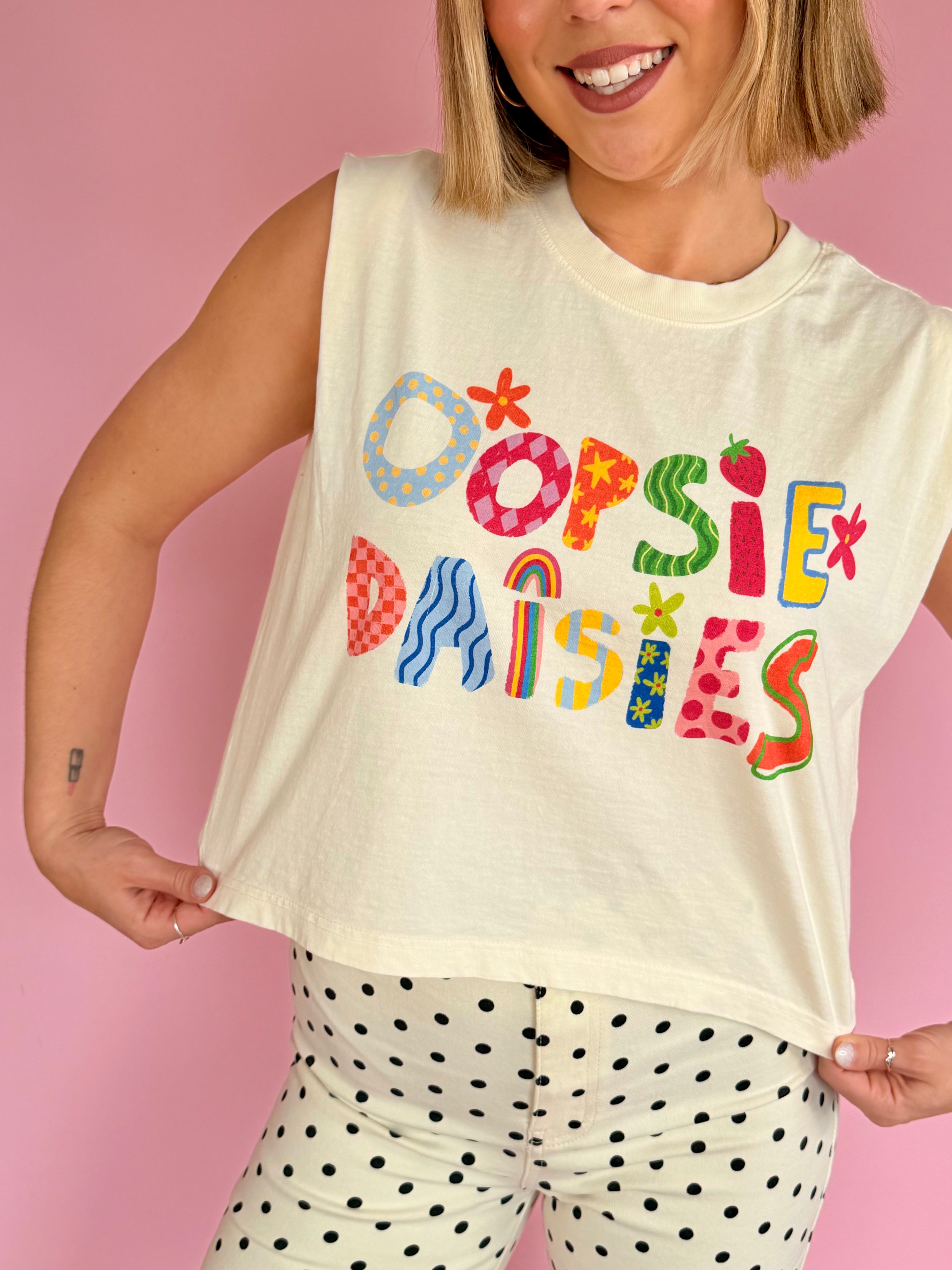 Person wearing a sleeveless shirt with colorful text 'OOPSIE DAISIES' against a pink background, pattern lover, sleeveless tee, flower and pattern contrast, cropped and flowy fit