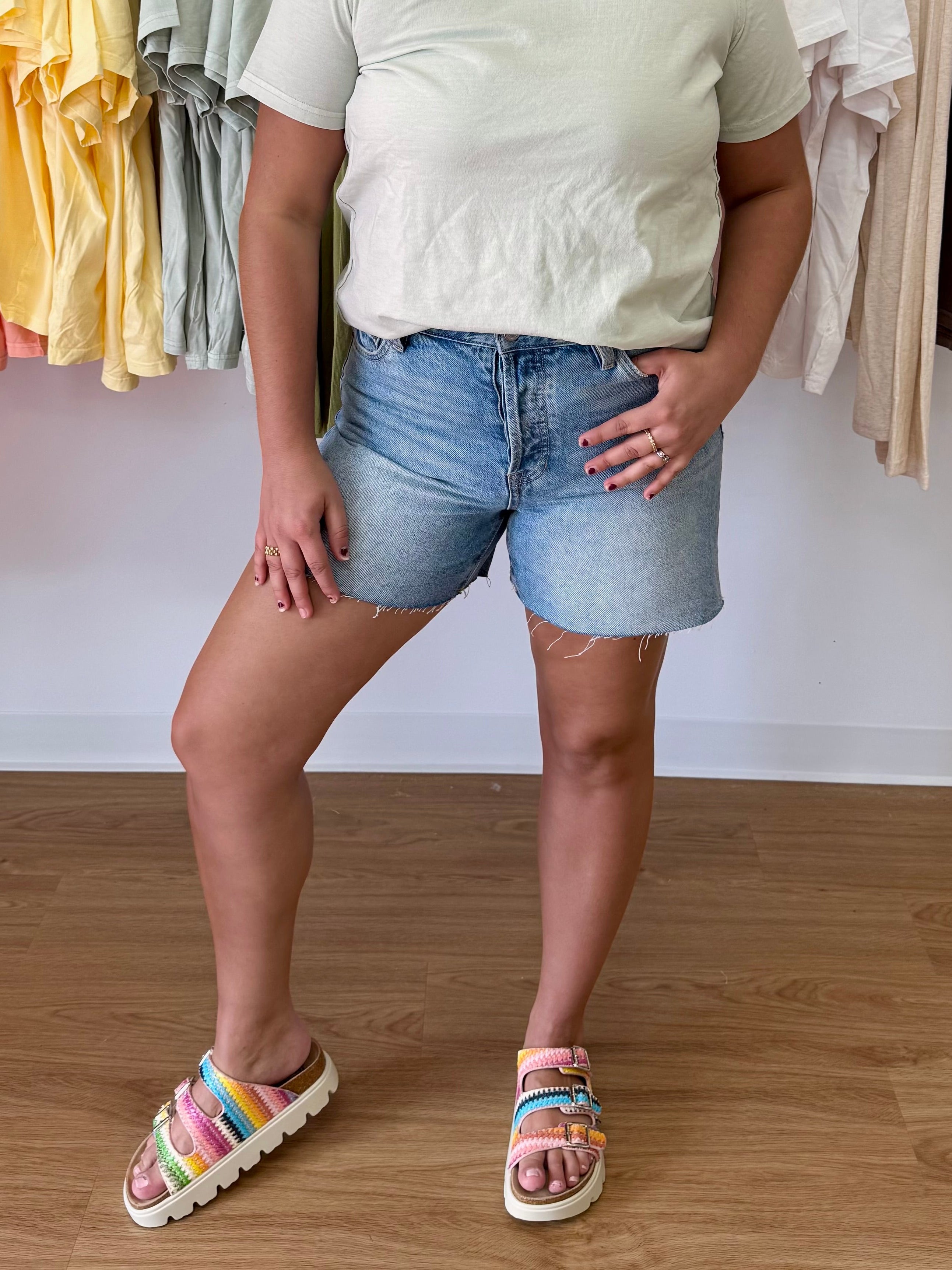 Person wearing a light-colored shirt and denim shorts standing on a wooden floor with clothing racks in the background, raw hem, short, button fly, stretchy