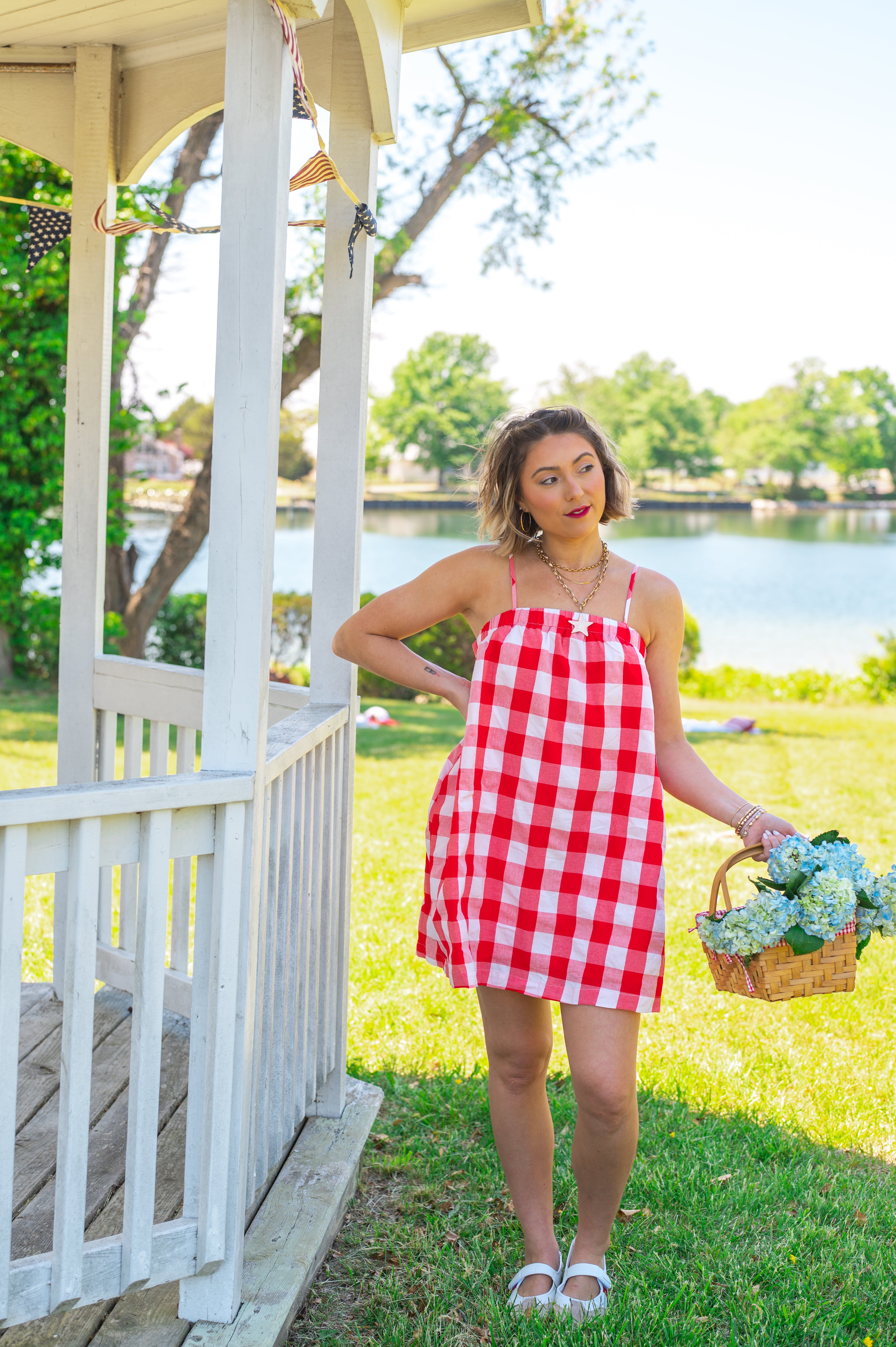 Woman in a red and white checkered dress standing by a gazebo with a lake in the background, red gingham, midi length, pockets, Americana, picnic cutie, flowy, straight fit, 