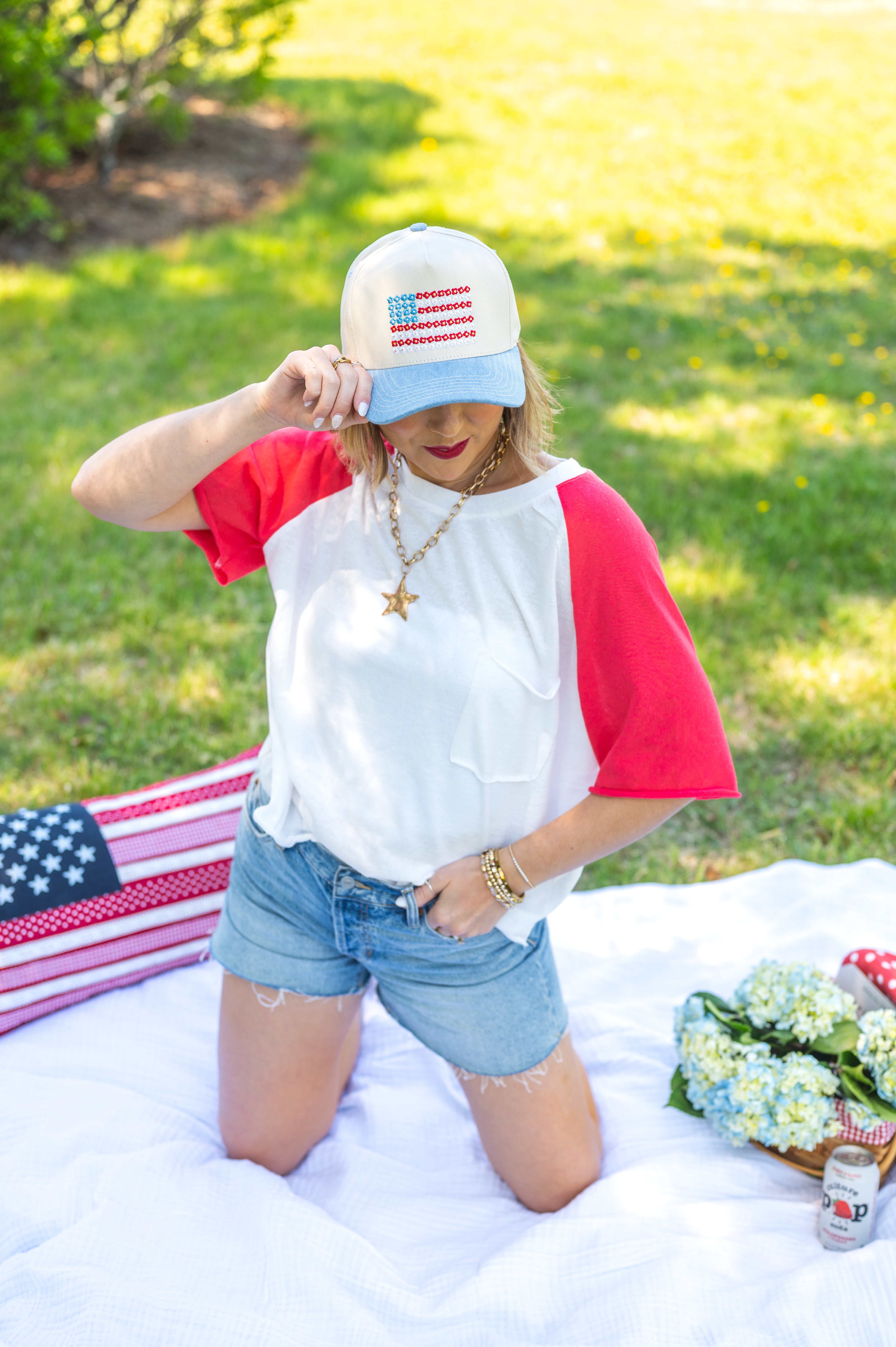 Woman in a red, white, and blue outfit with an American flag hat on a grassy outdoor setting, loose-fit tee shirt, red and white, USA, airy, breathable, pocket tee, raw hem , red sleeve