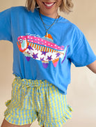 Person wearing a blue t-shirt with a colorful fish design and yellow checkered shorts on a beige background, patchwork tee, stars and polka dot, fish graphic tee, summer block color tee 