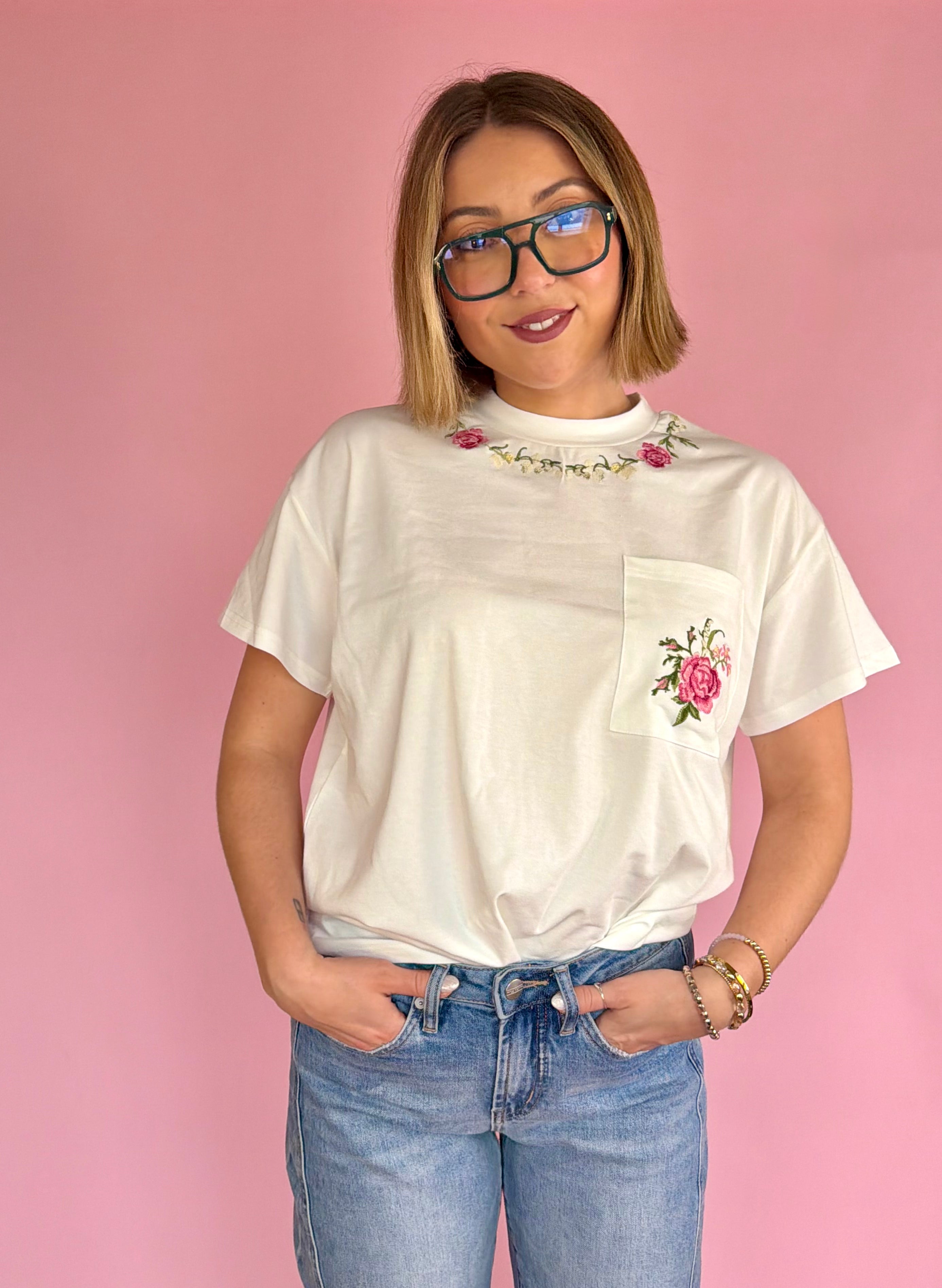 Woman wearing a white t-shirt with floral designs on a pink background, flower neckline, flower pocket tee, full length white tee, comfy and casual, effortless dressy tee 