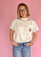 Woman wearing a white t-shirt with floral designs on a pink background, flower neckline, flower pocket tee, full length white tee, comfy and casual, effortless dressy tee 