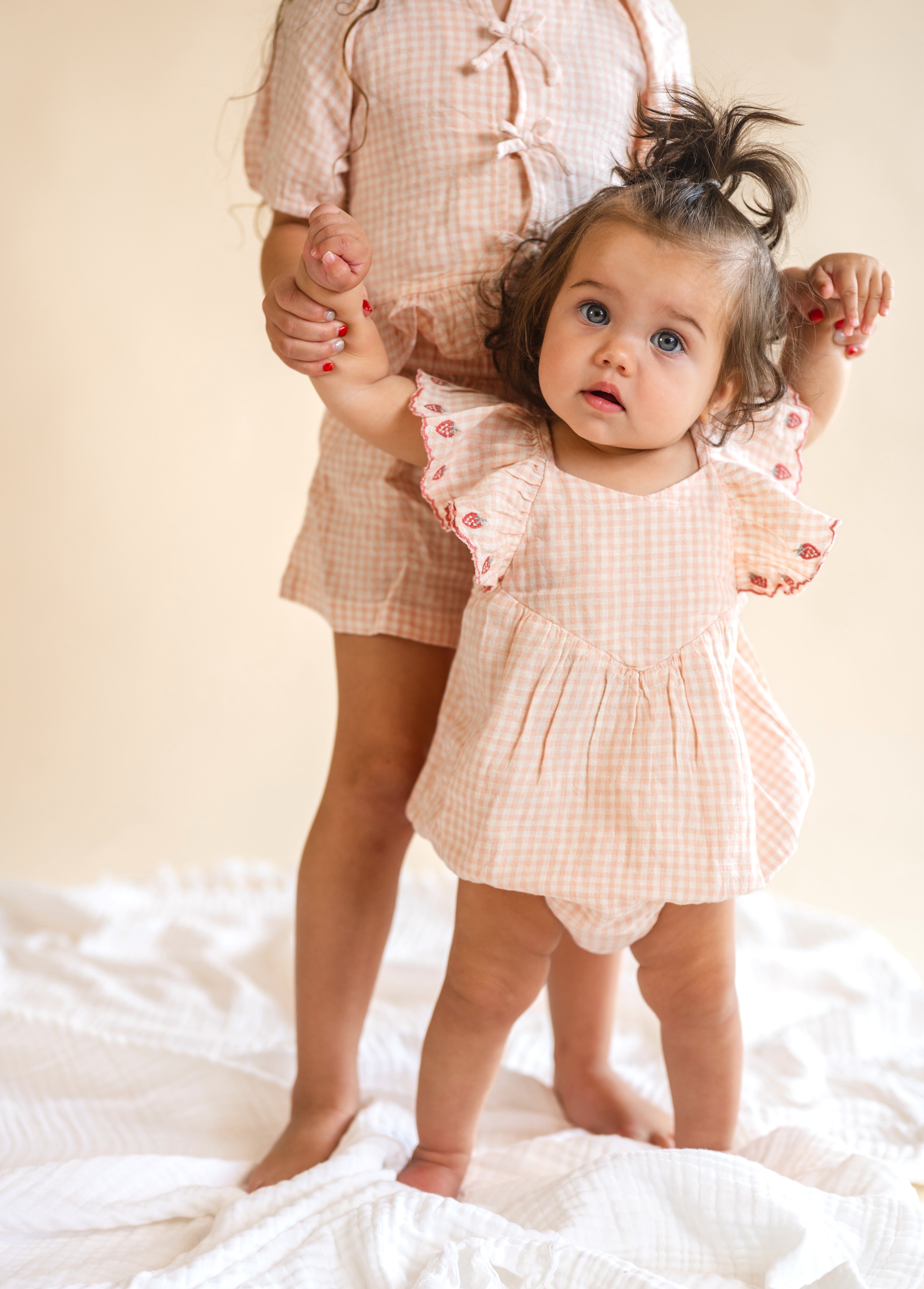 Child in a matching checkered outfit standing on a white surface with a neutral background, strawberry lined sleeves, flowy romper, baby wear, pink plaid