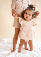 Child in a matching checkered outfit standing on a white surface with a neutral background, strawberry lined sleeves, flowy romper, baby wear, pink plaid