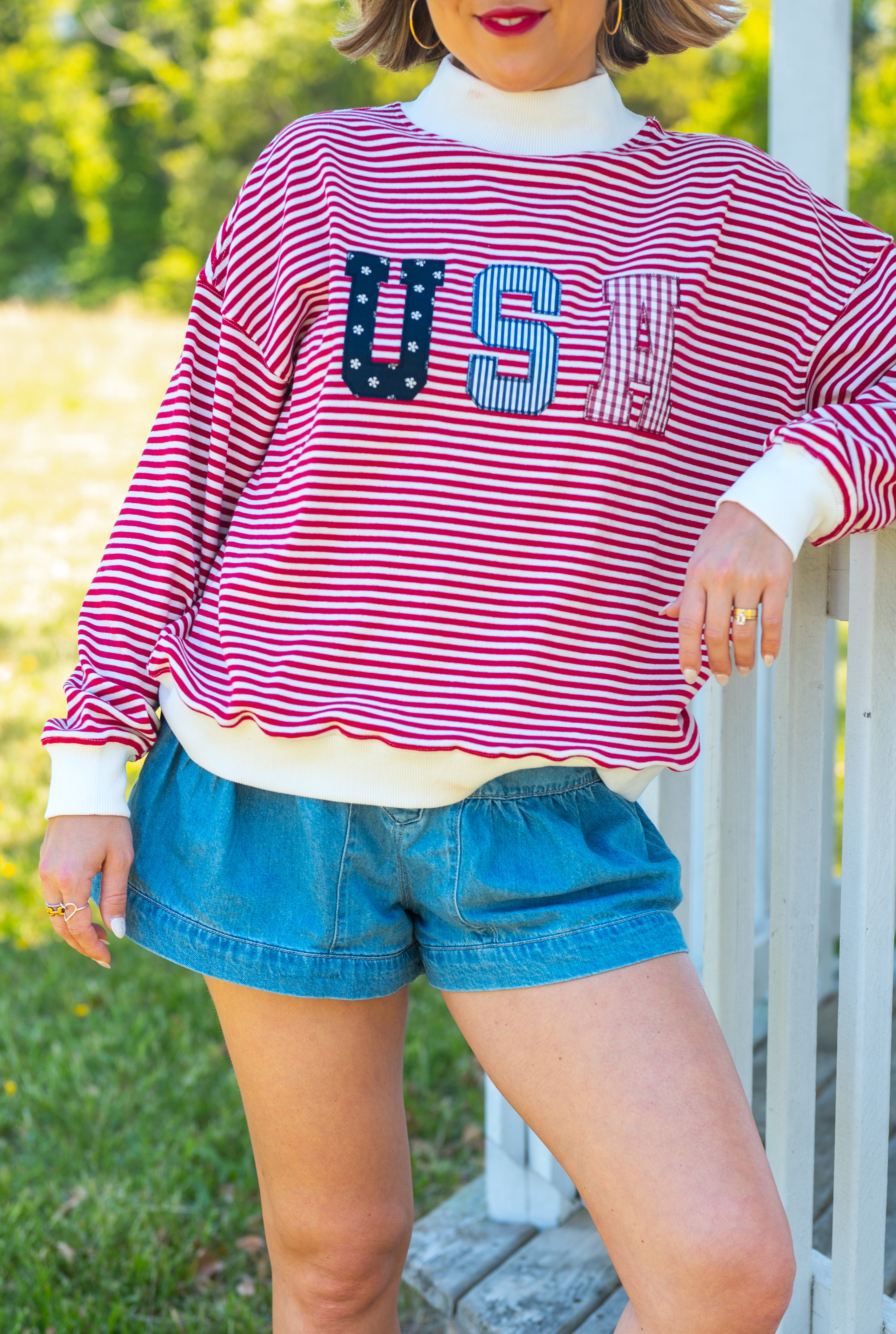 Person wearing a red and white striped shirt with 'USA' print, denim shorts, and white sneakers outdoors, USA, americana, striped, patchwork