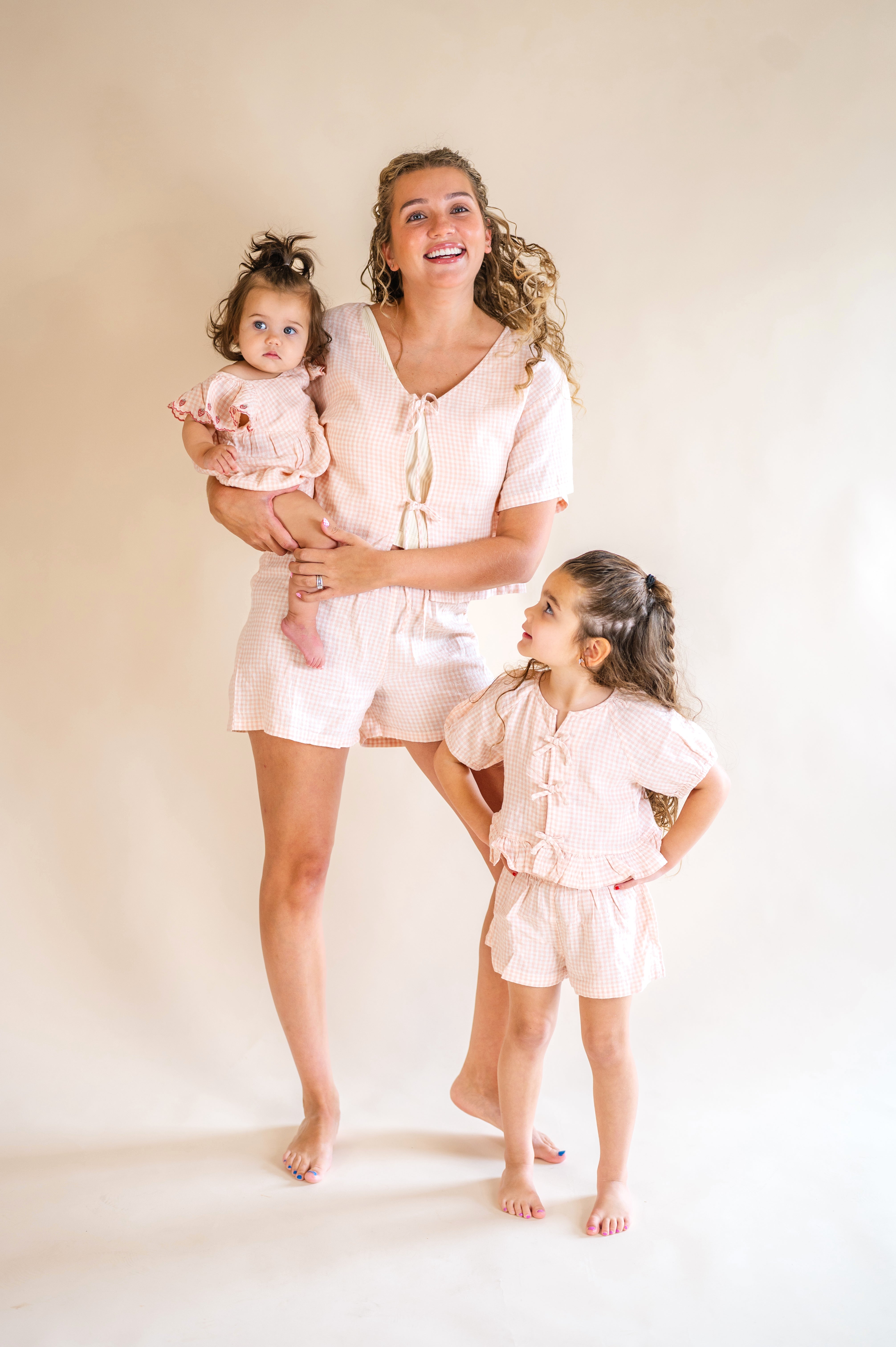 Woman and two young girls in matching light pink outfits standing against a plain background, pink plaid set, mommy and me, tie front top, plaid short set, pink and white matching set