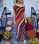 Model wearing a one-shoulder maxi dress in diagonal chocolate, rust, cream, and gold stripes with a draped cowl neckline and a fitted floor-length silhouette.